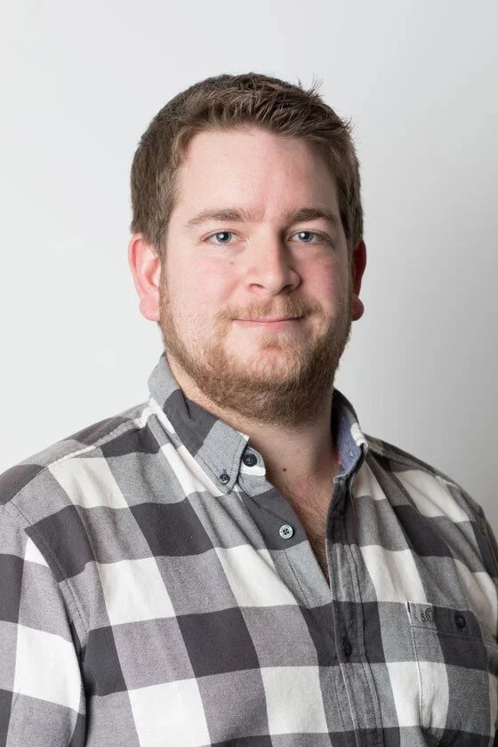 Porträt eines jungen Mannes mit braunen Haaren, Vollbart und blauäugigen Blick, trägt ein graues kariertes Hemd, vor hellem Hintergrund.