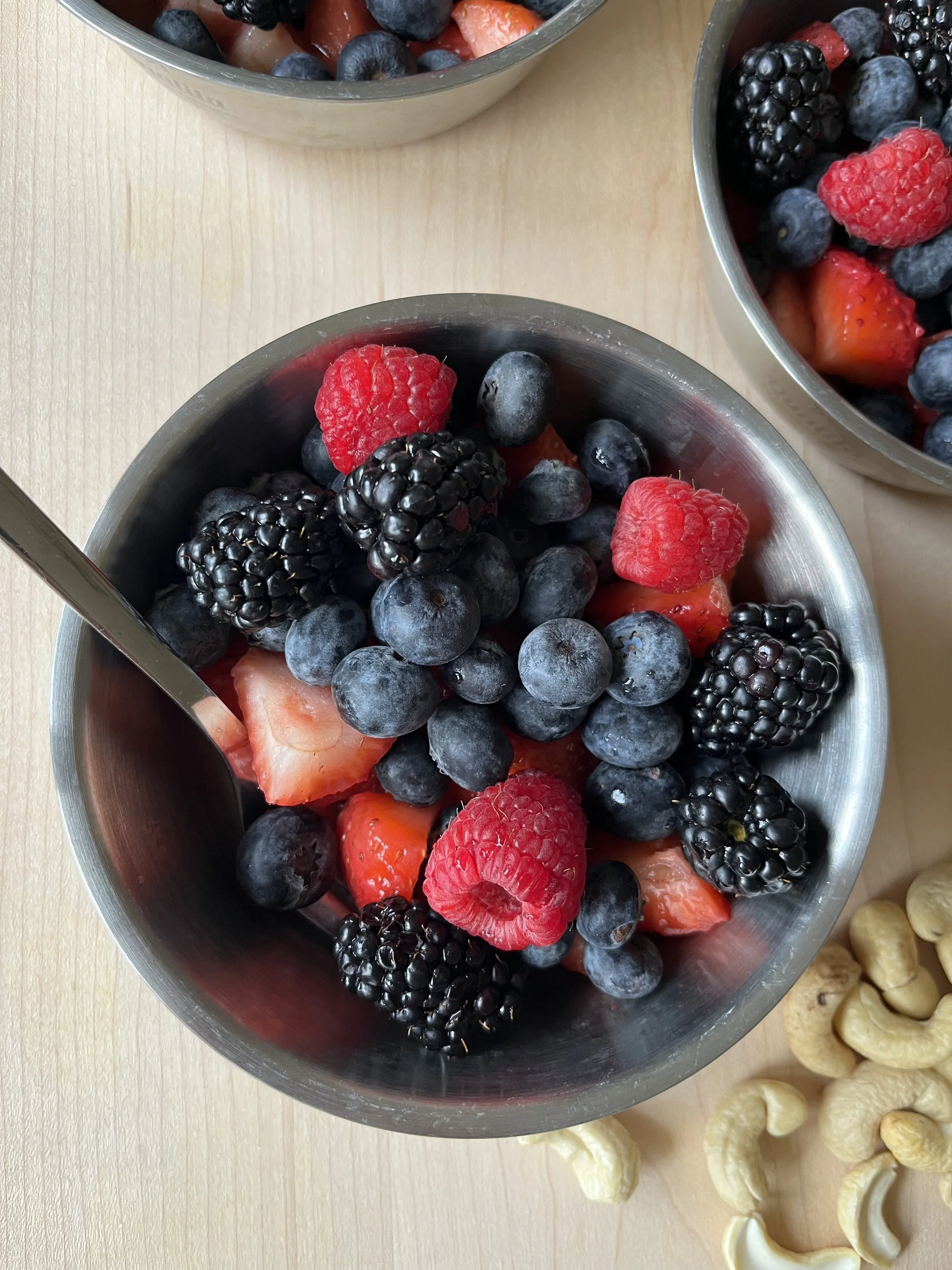 A bowl of mixed fresh berries including strawberries, blueberries, blackberries, and raspberries on a light wooden surface, with another similar bowl partially visible in the background and some cashew nuts nearby.