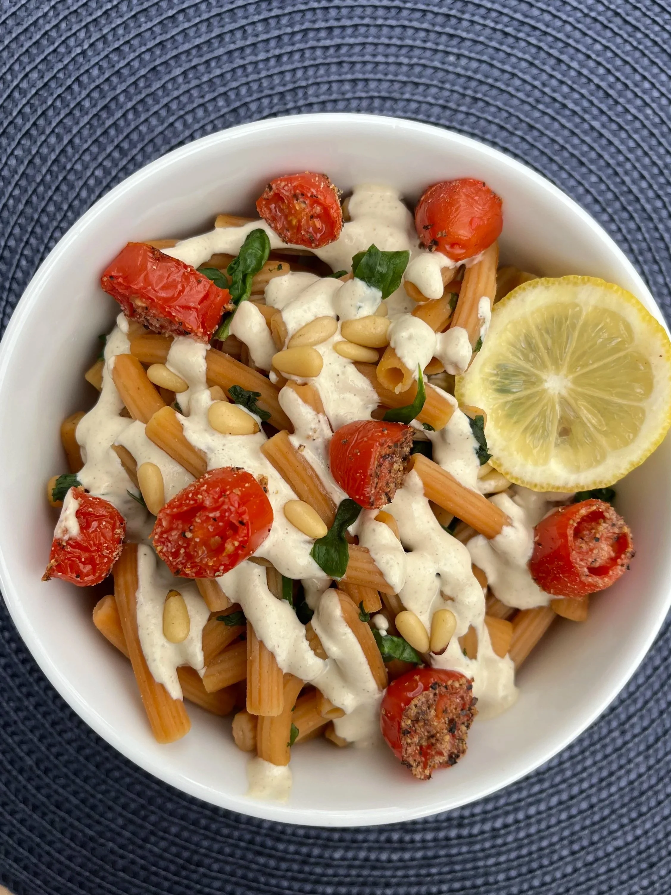 Bowl of cooked pasta with cherry tomatoes, pine nuts, chopped herbs, and a lemon wedge, topped with a creamy white sauce.