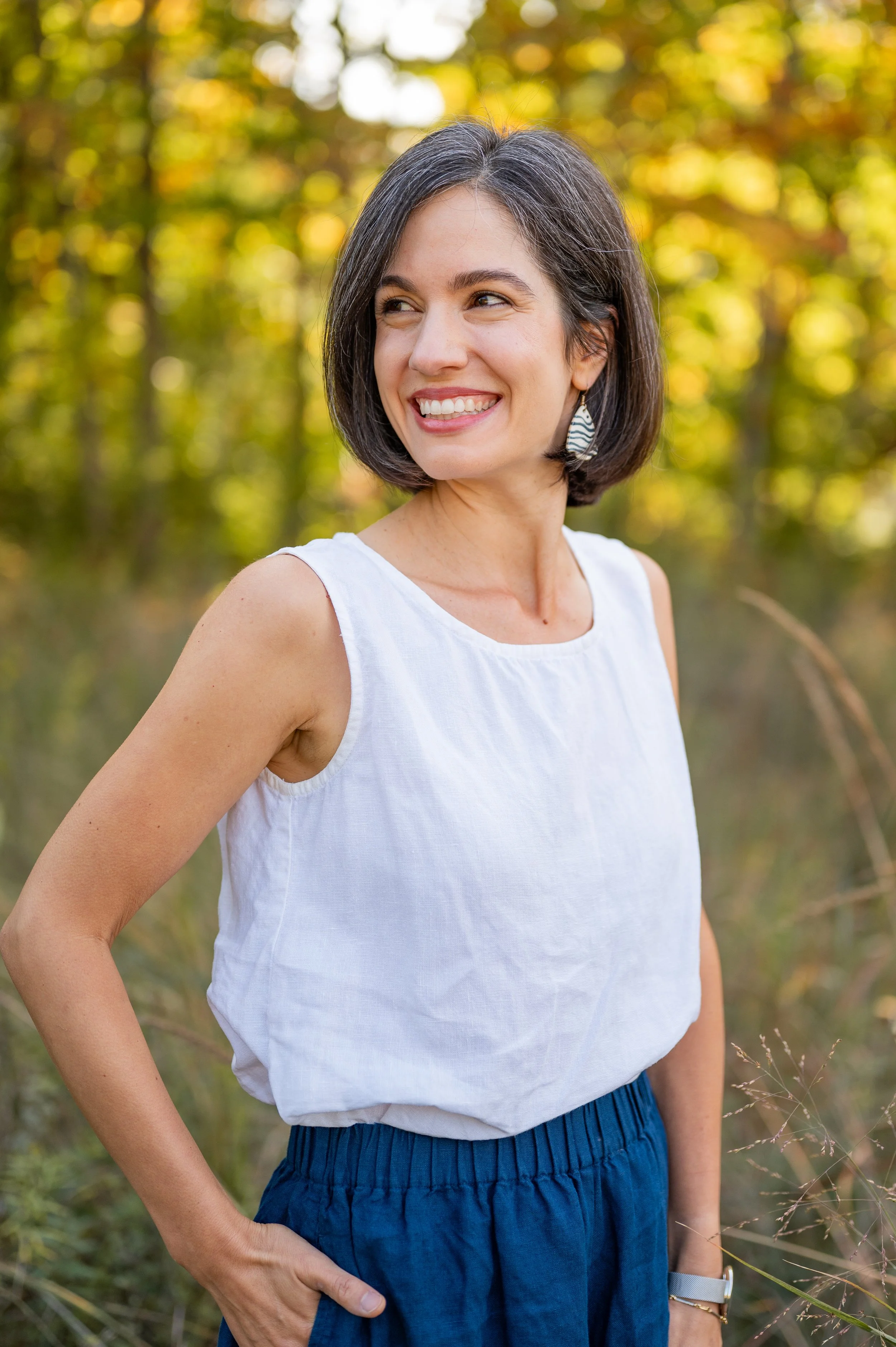 A woman smiling outdoors wearing a white sleeveless top and dark blue pants, with a blurred background of trees and green foliage.