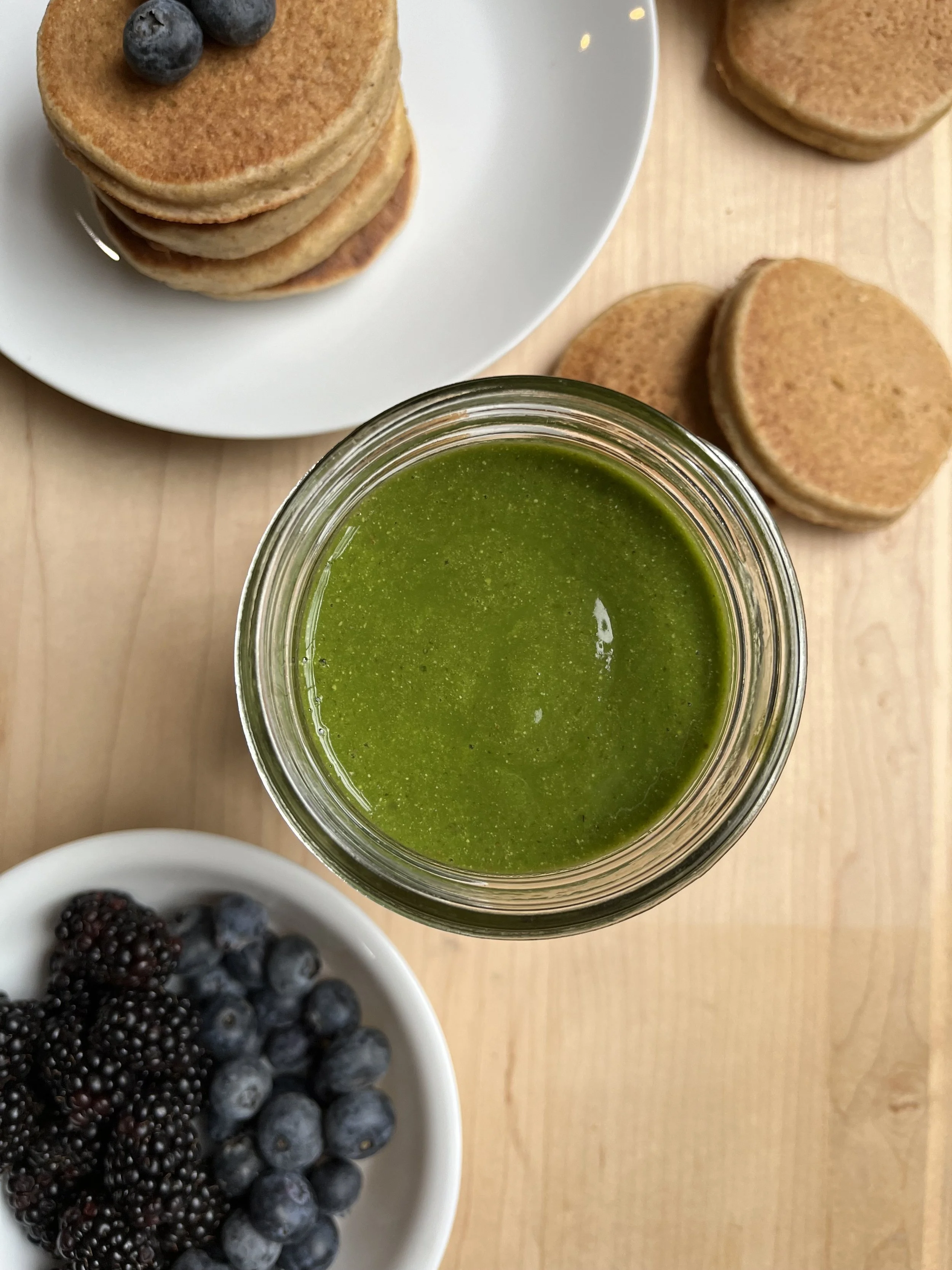 Stack of four pancakes topped with blueberries on a white plate, a jar of green smoothie or juice, a bowl of mixed blackberries and blueberries, and additional pancakes on a wooden surface.
