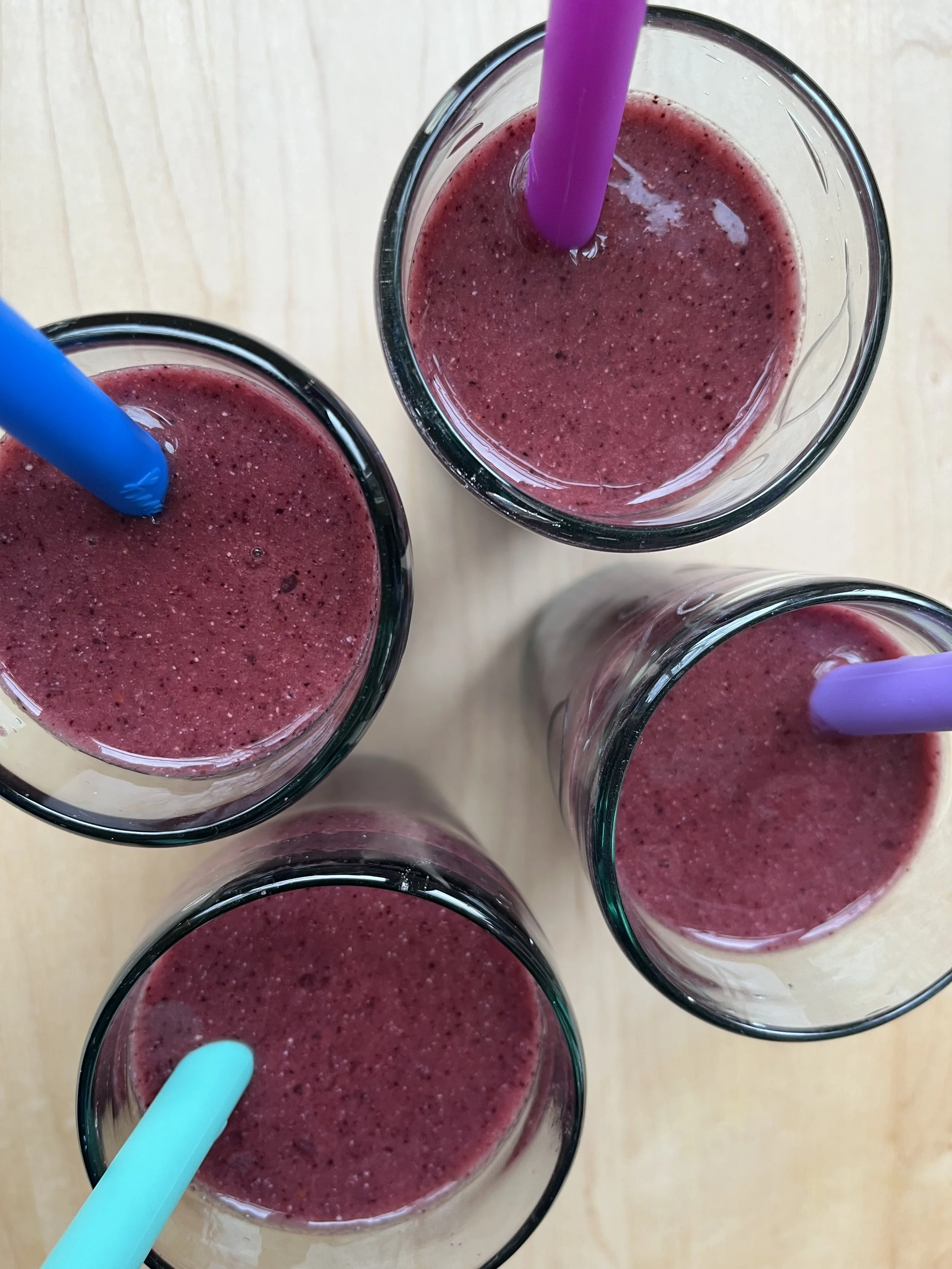 Five glasses of berry smoothies with colorful straws on a light wooden surface.