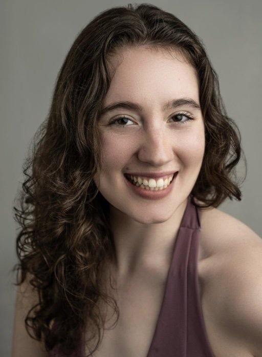 Young woman with wavy brown hair smiling, wearing a purple sleeveless top, against a neutral background.