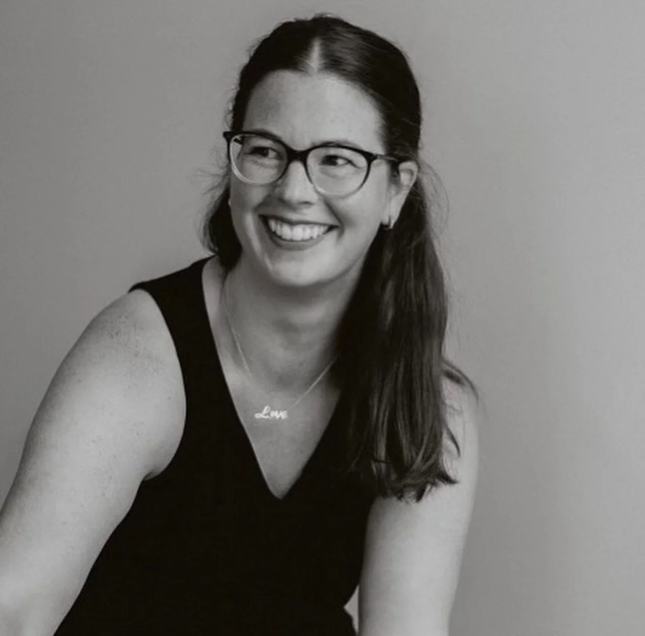 Black and white photo of a woman with glasses smiling, wearing a necklace with the word 'Love.'