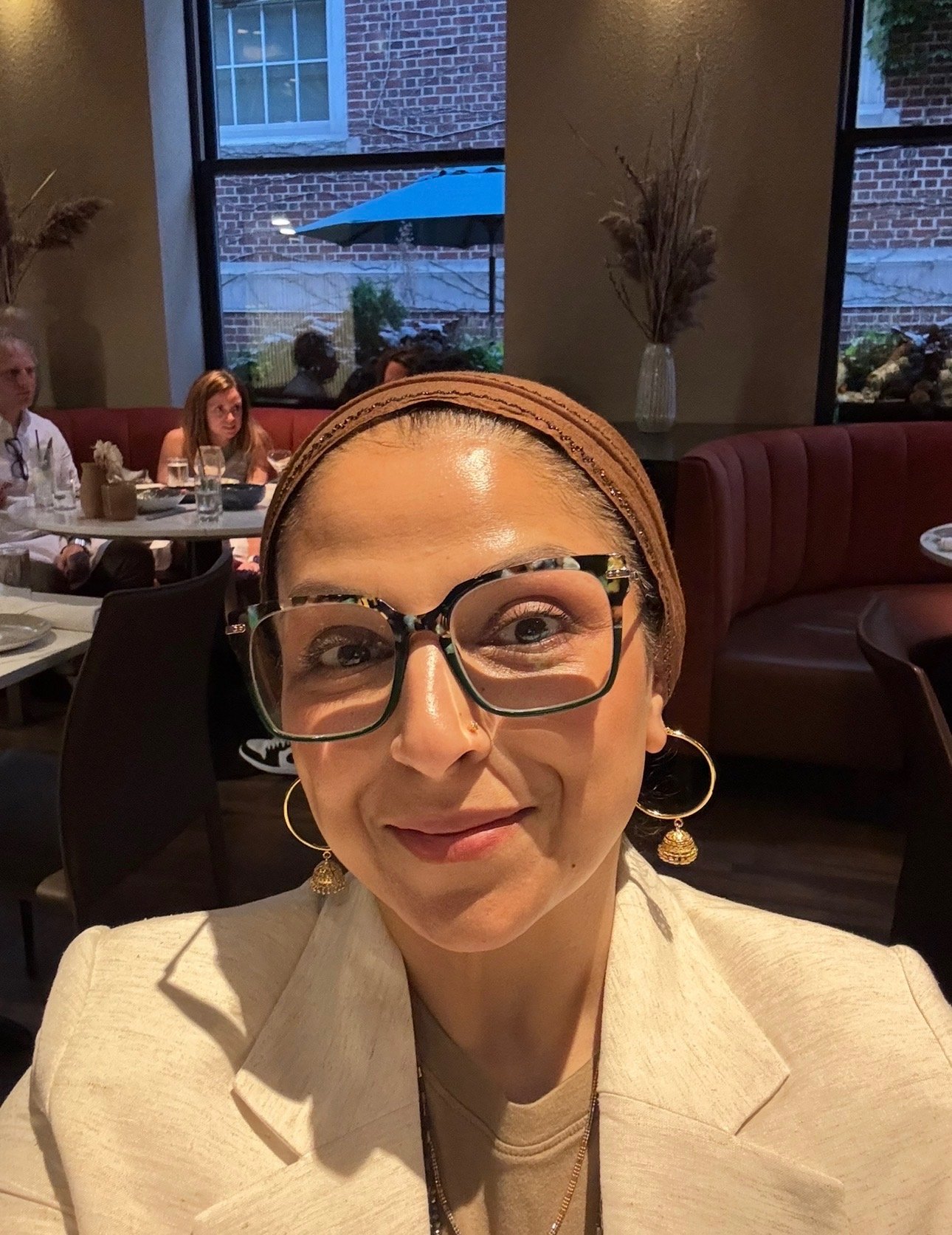 Close-up of a woman wearing glasses, earrings, and a headband, sitting in a restaurant with a group of people in the background.