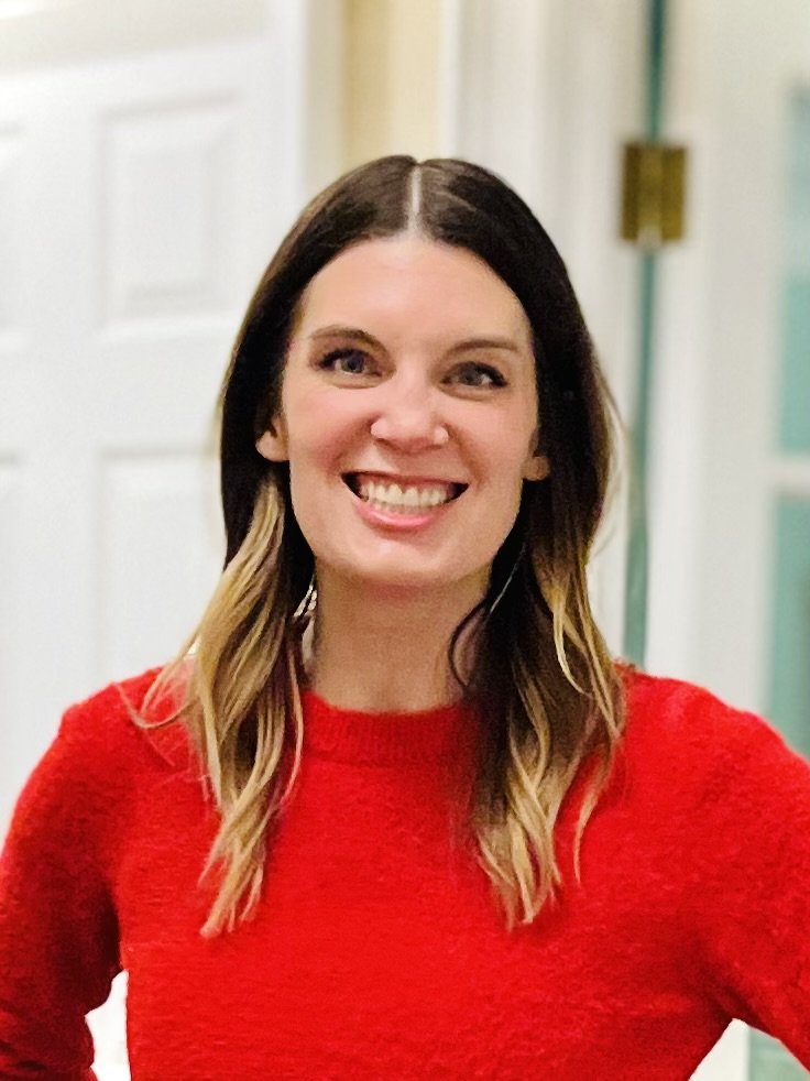 A woman with brown hair and blonde highlights smiling and wearing a red sweater indoors.