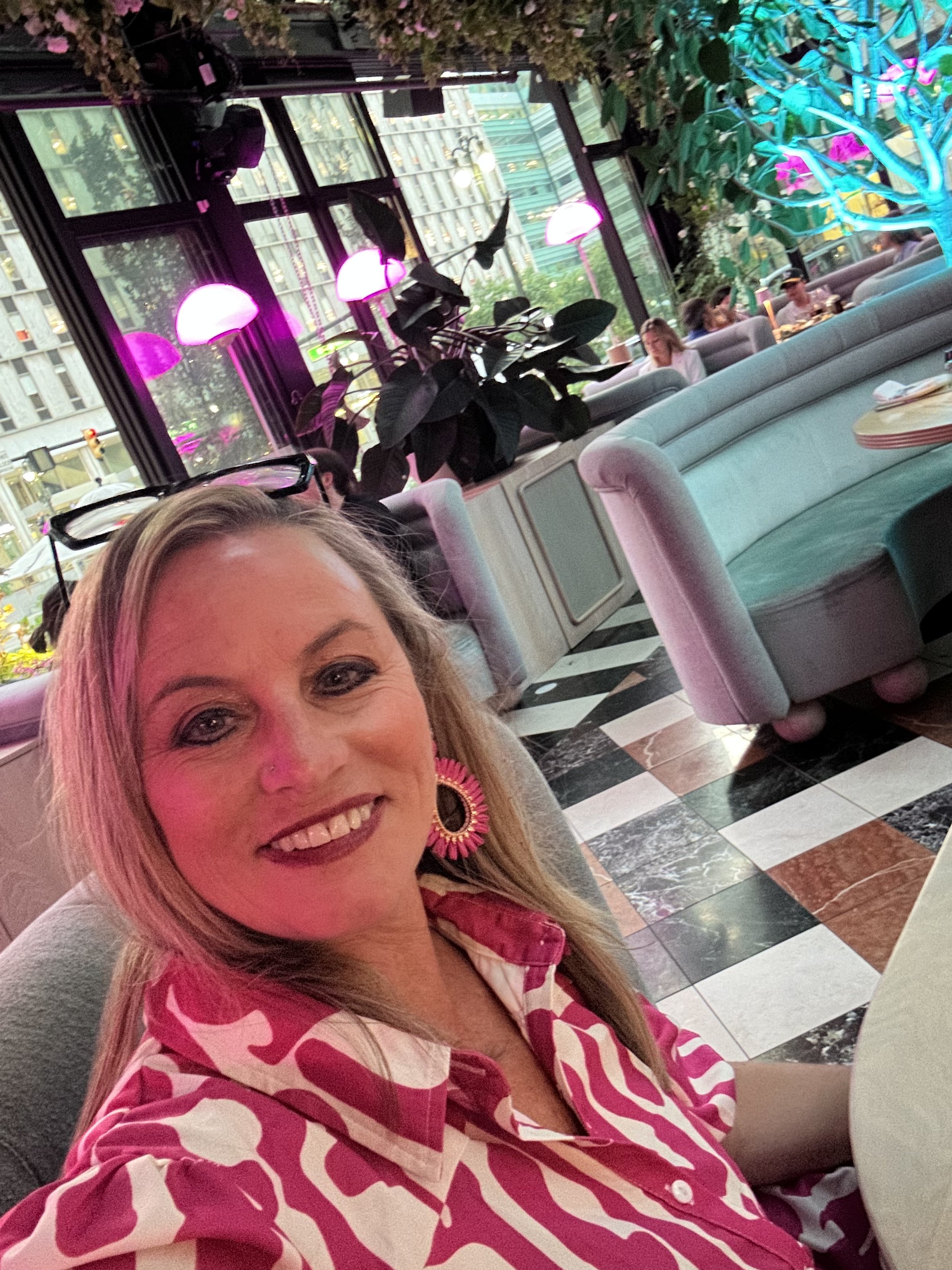 A woman with blonde hair smiling at the camera in a modern, colorful restaurant with checkered black-and-white and brown marble flooring, velvet seating, large windows, and pink neon lighting. Other patrons are visible sitting at tables in the background.