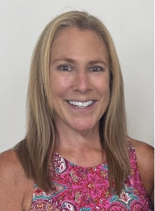 A woman with shoulder-length blonde hair smiling at the camera, wearing a sleeveless pink, purple, and blue paisley patterned top against a plain white background.