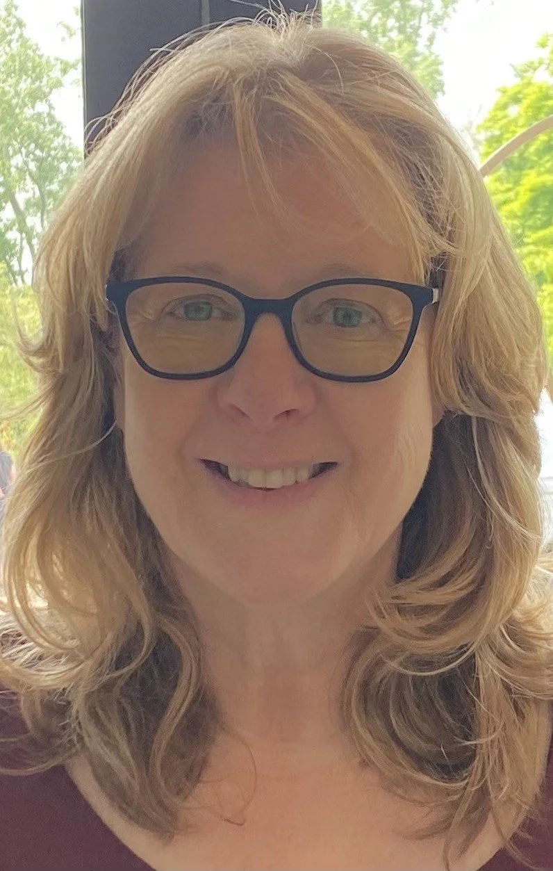 A woman with blonde, wavy hair and glasses smiling, with greenery visible through a window behind her.