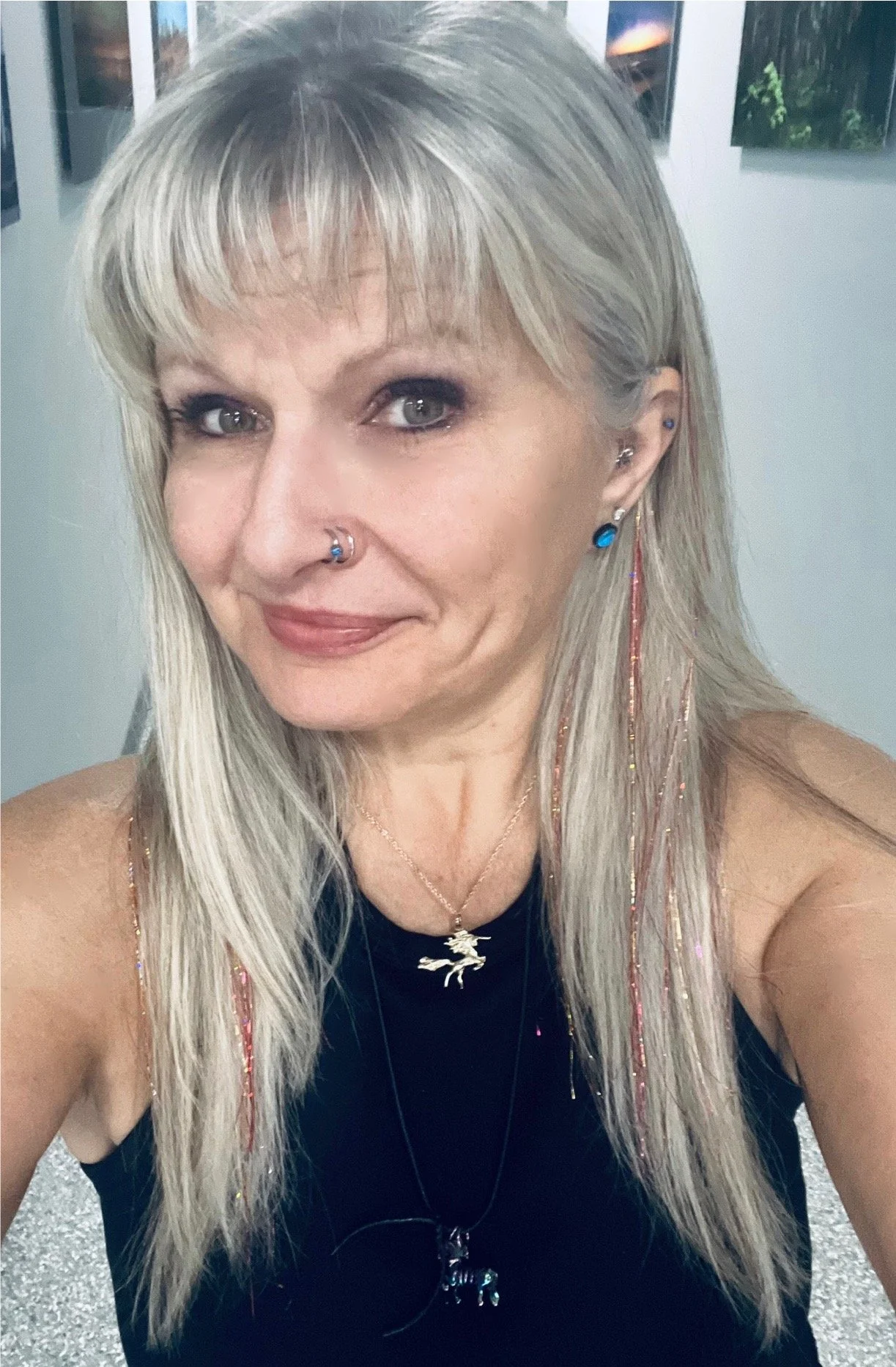 A woman with long blonde hair, blue eyes, and multiple earrings, including a septum piercing, smiling at the camera, wearing a black sleeveless top and jewelry including necklaces and earrings, in an art gallery with framed pictures on the wall.