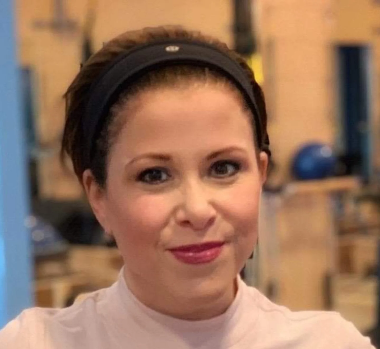 Portrait of a woman with short dark hair, wearing a beige top and a black headband, smiling in a gym setting.