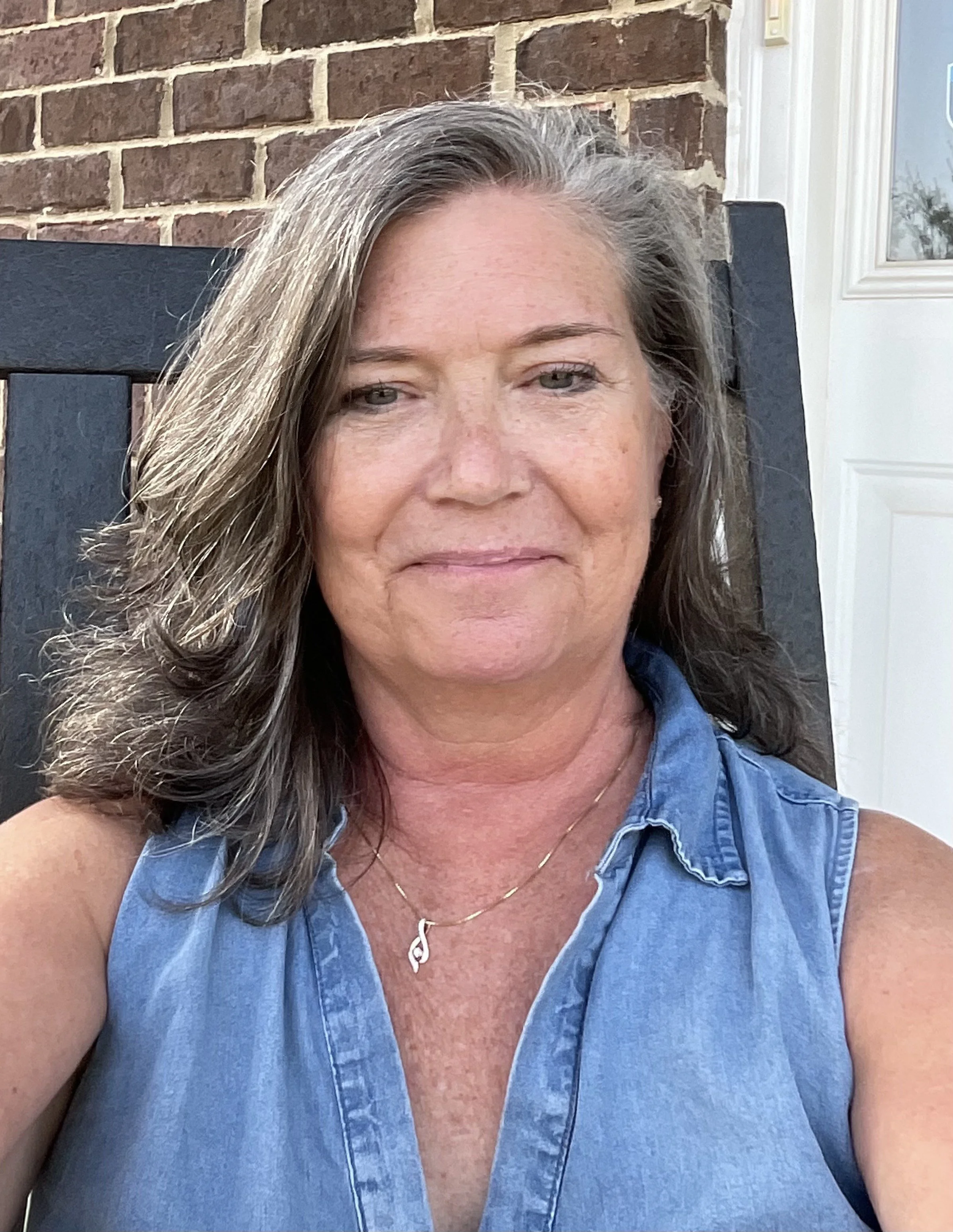 A woman with gray, wavy hair sitting outdoors on a black chair, wearing a sleeveless denim shirt and a gold necklace with a harp pendant, in front of a brick wall and a white-framed window.