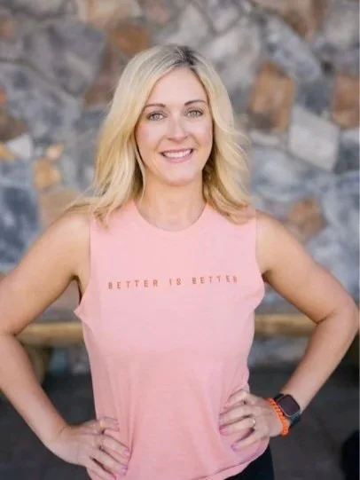 A smiling woman with blonde hair, wearing a pink sleeveless top that says 'Better is Better,' standing in front of a stone wall.
