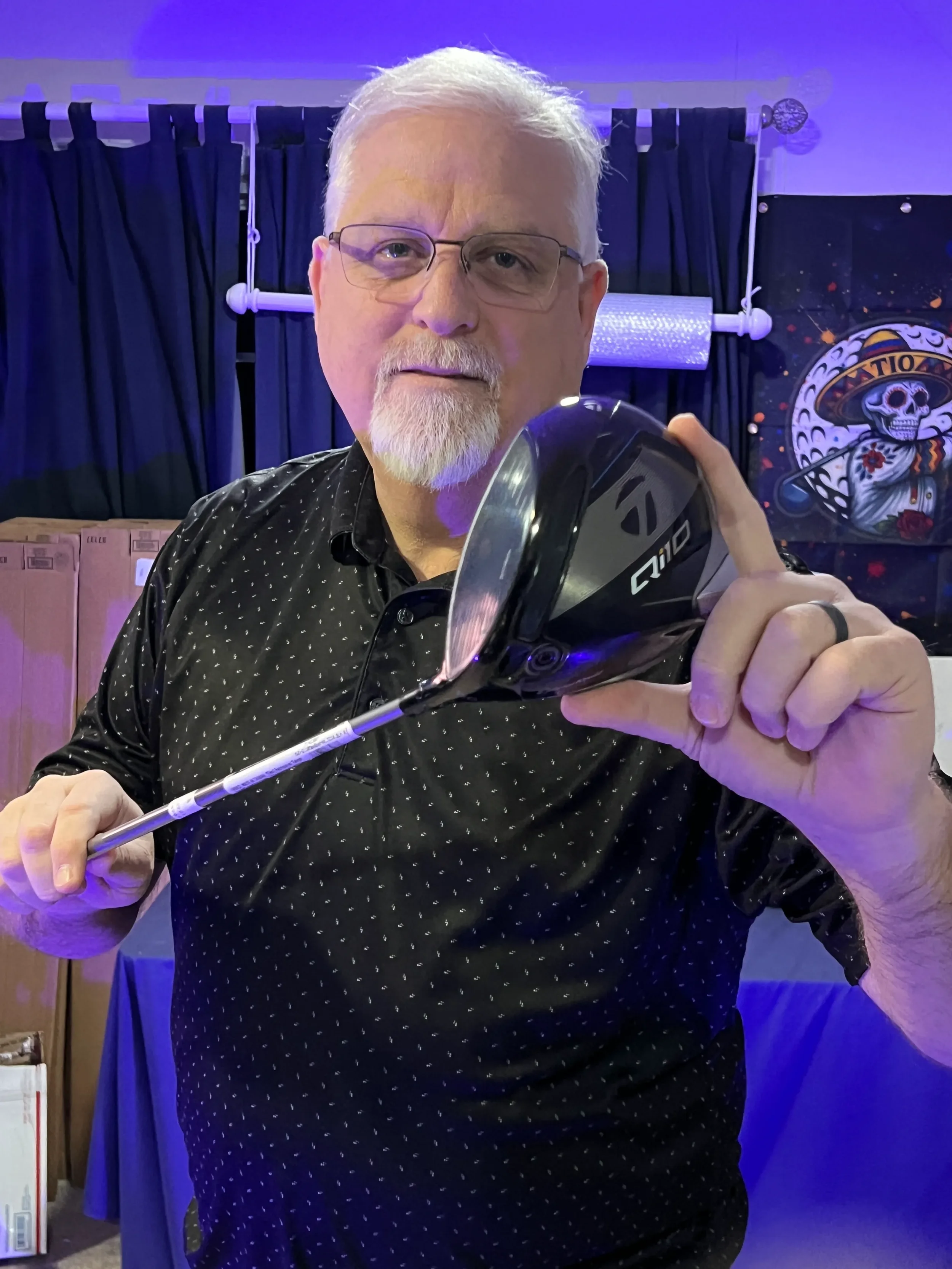 Uncle Steve holding a golf driver indoors with a dark blue curtain and a colorful celestial-themed artwork in the background.