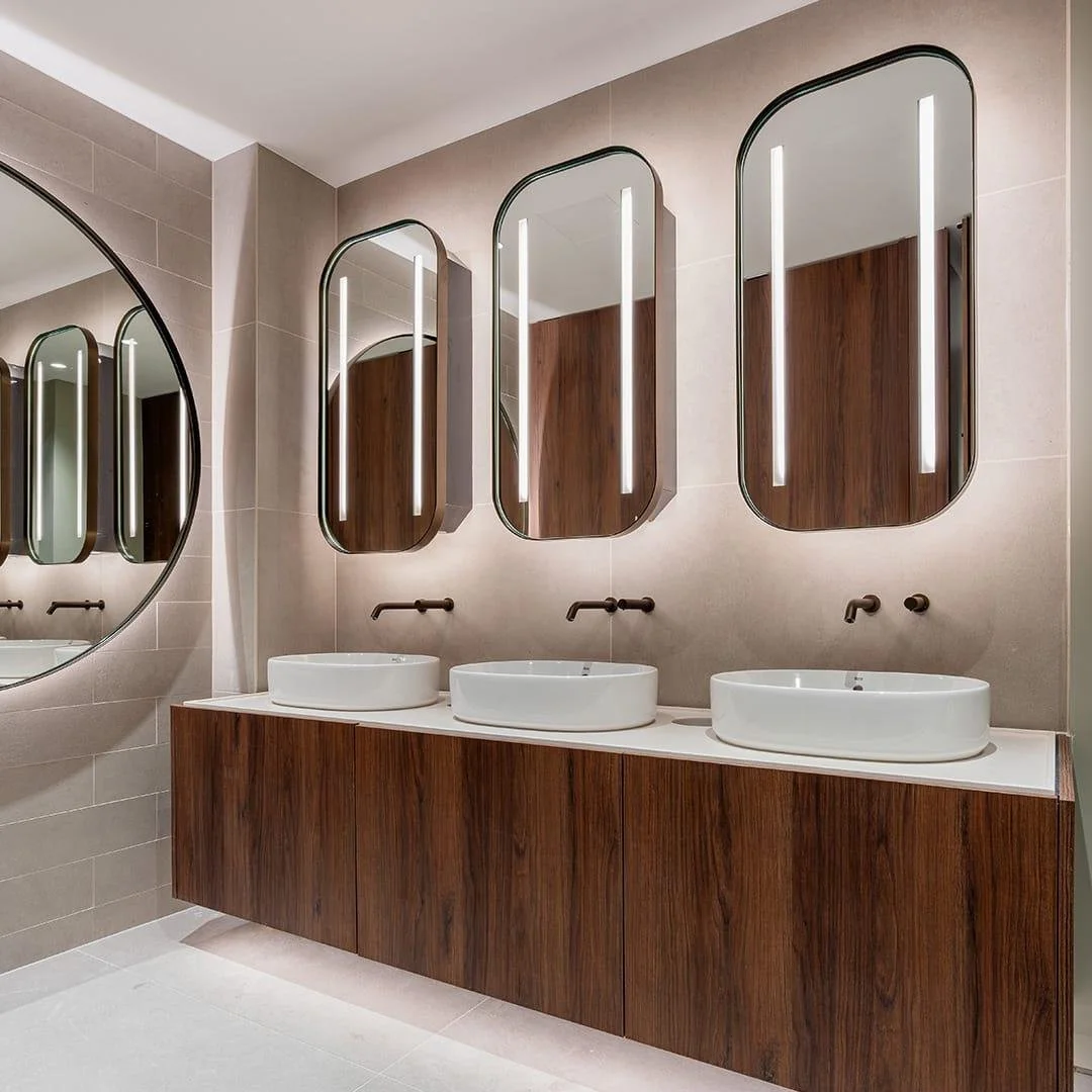 Modern bathroom vanity with three white vessel sinks, wood cabinetry, illuminated arched mirrors, and wall-mounted black faucets