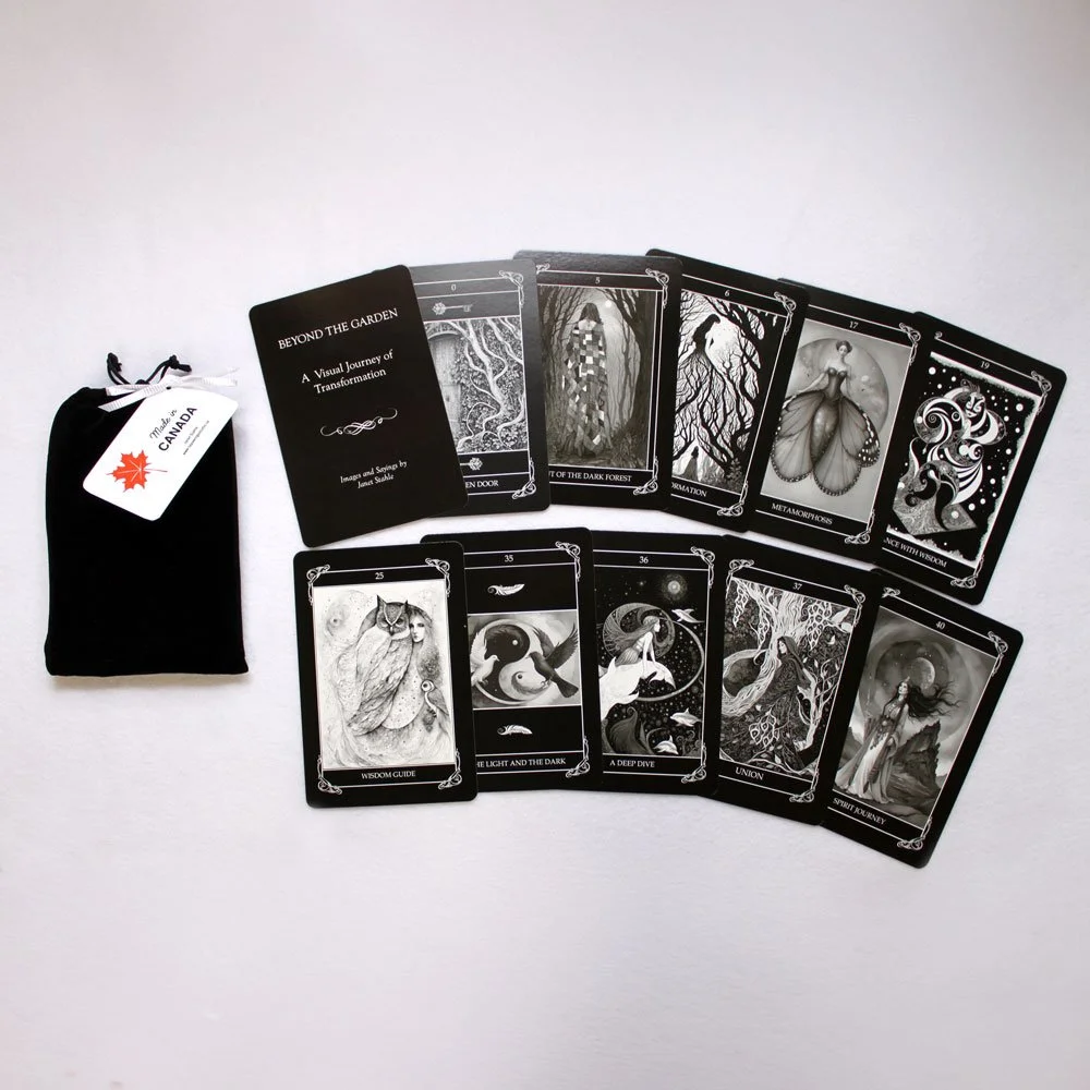 A set of black tarot or oracle cards with detailed artwork, some face up displaying different symbols and images, laid out on a white surface. There is a small black cloth bag with a white tag that reads 'Made in Canada' to the left of the cards.