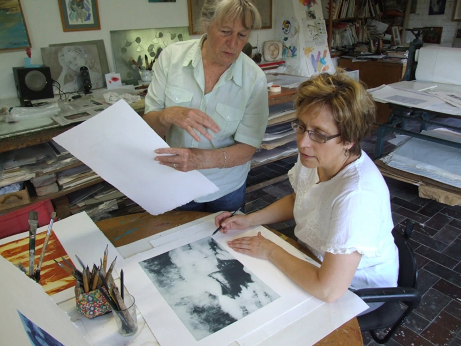 An artist and a woman examining artwork in an art studio filled with papers, paintings, and art supplies.