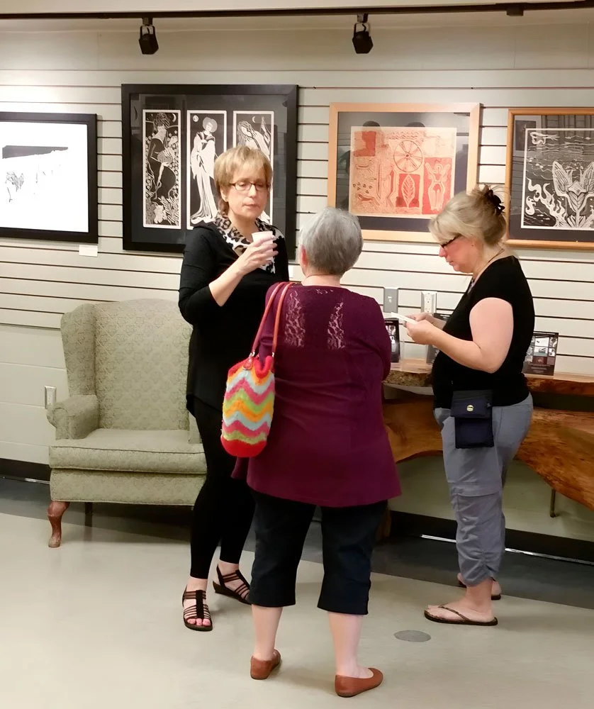 Three women standing in an art gallery, viewing framed artwork on the wall. One woman is holding a drink, another is looking at her phone, and the third, with gray hair, has her back to the camera.