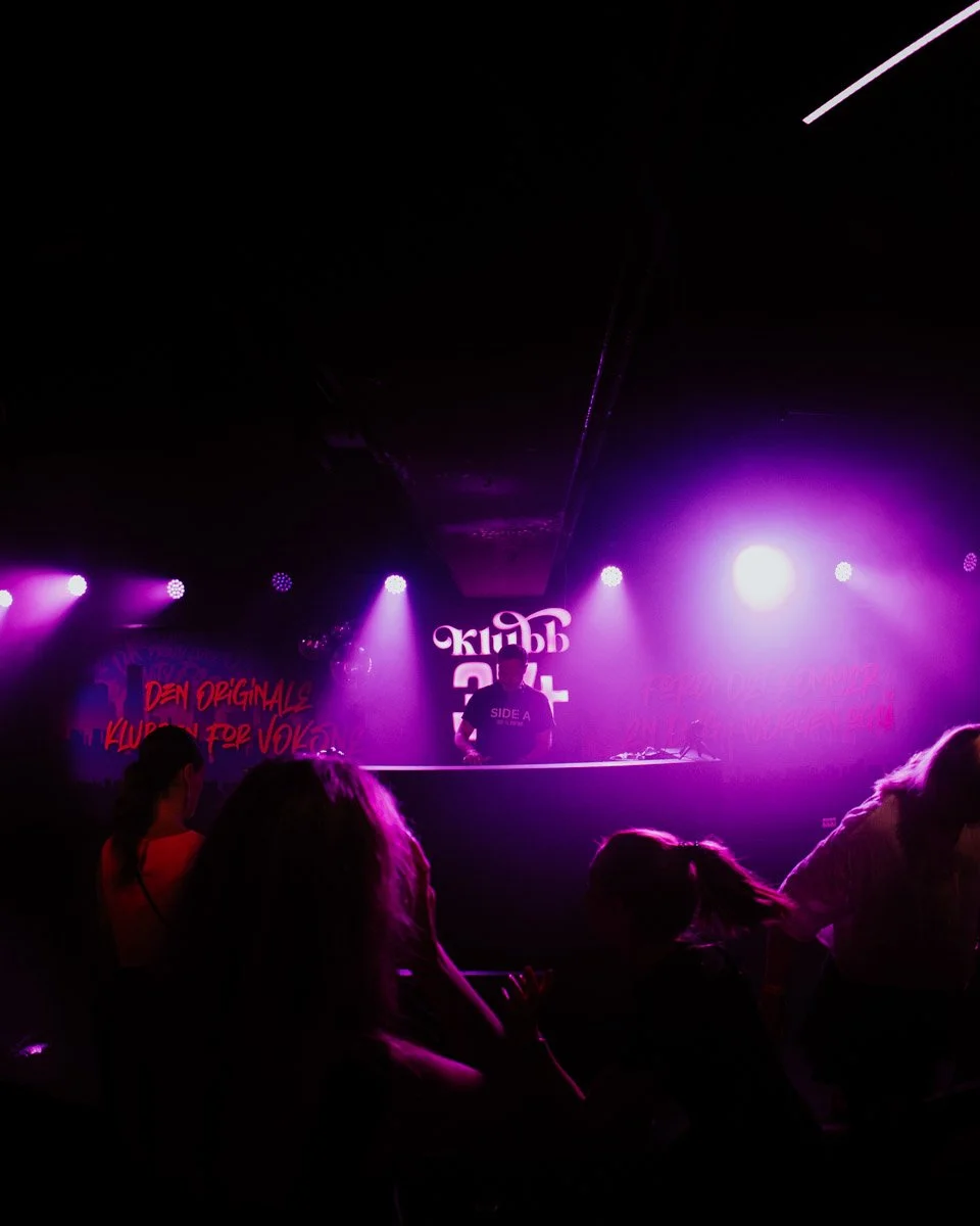 En klubbscene med en DJ på scenen og flere mennesker som danser. Mørkt rom med lilla og hvite lys. Vegg med tekster, blant annet "DEN ORIGINALE KLUBBEN FOR VOX".