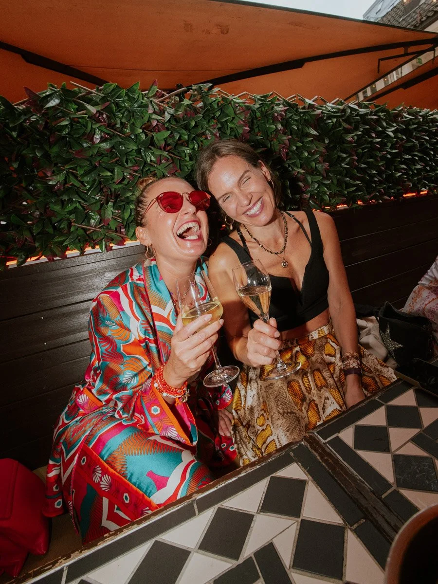 To voksne kvinner smiler og ler, de holder champagneglass og koser seg på en restaurant. En av kvinnene har på seg rød solbriller og fargerik klær, den andre har på seg en svart topp og skinnende skjørt. De sitter ved et bord med sort-hvite fliser.