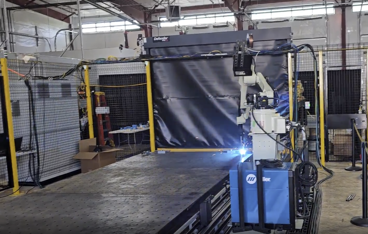A robotic welding arm in a factory, welding metal sheets on a large worktable, with sparks visible at the welding point.