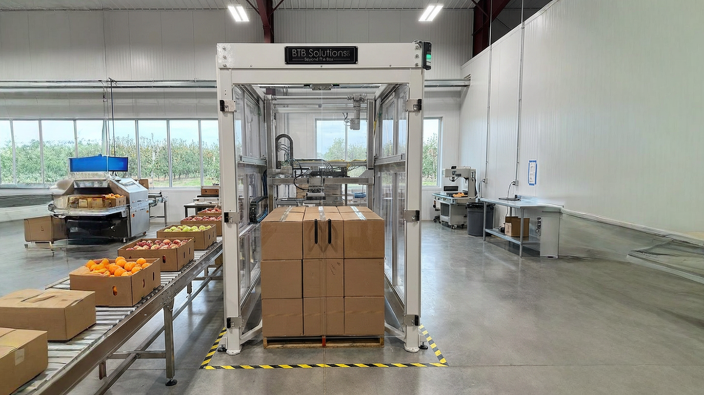 A manufacturing or packaging facility with a robotic system labeled 'BTB Solutions' handling boxes, with fruits like oranges and apples in cardboard boxes and on roller conveyor belts, and various machines and workstations in a spacious, well-lit room.