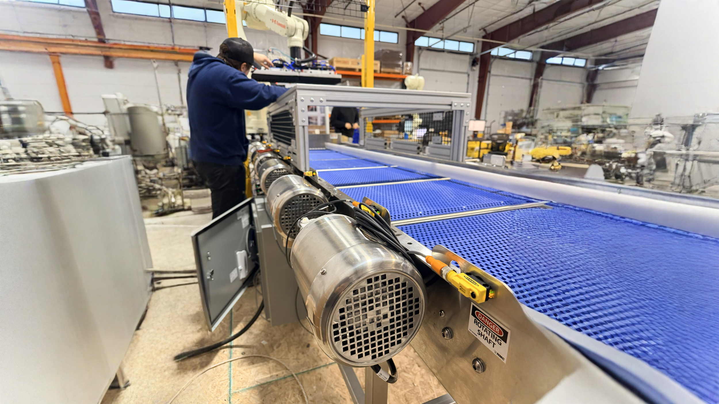 Worker in a blue hoodie and cap working on a large industrial machine with blue textured surface in a spacious warehouse.