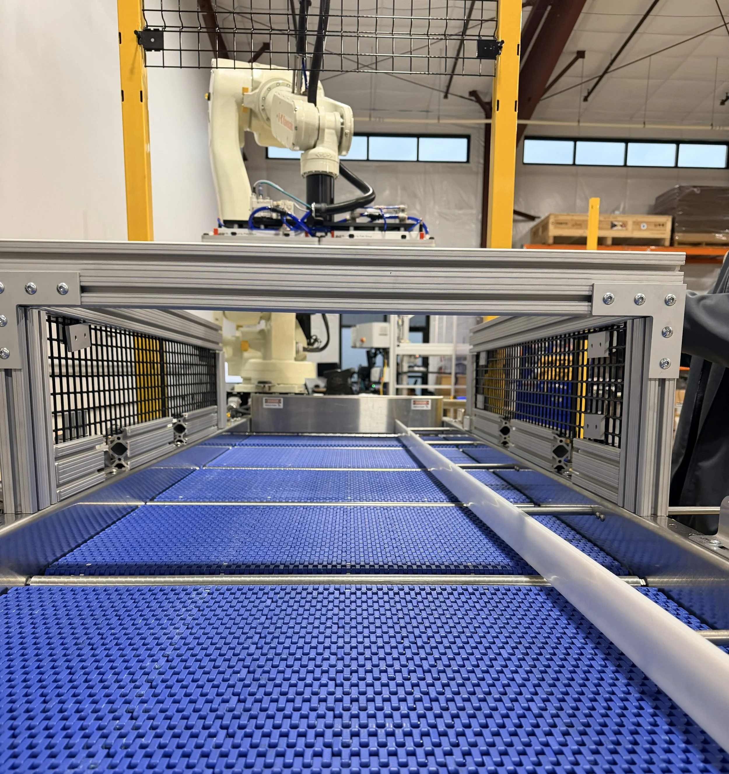View of an industrial robotic arm operating inside a metal frame with a blue conveyor belt, in a manufacturing facility.
