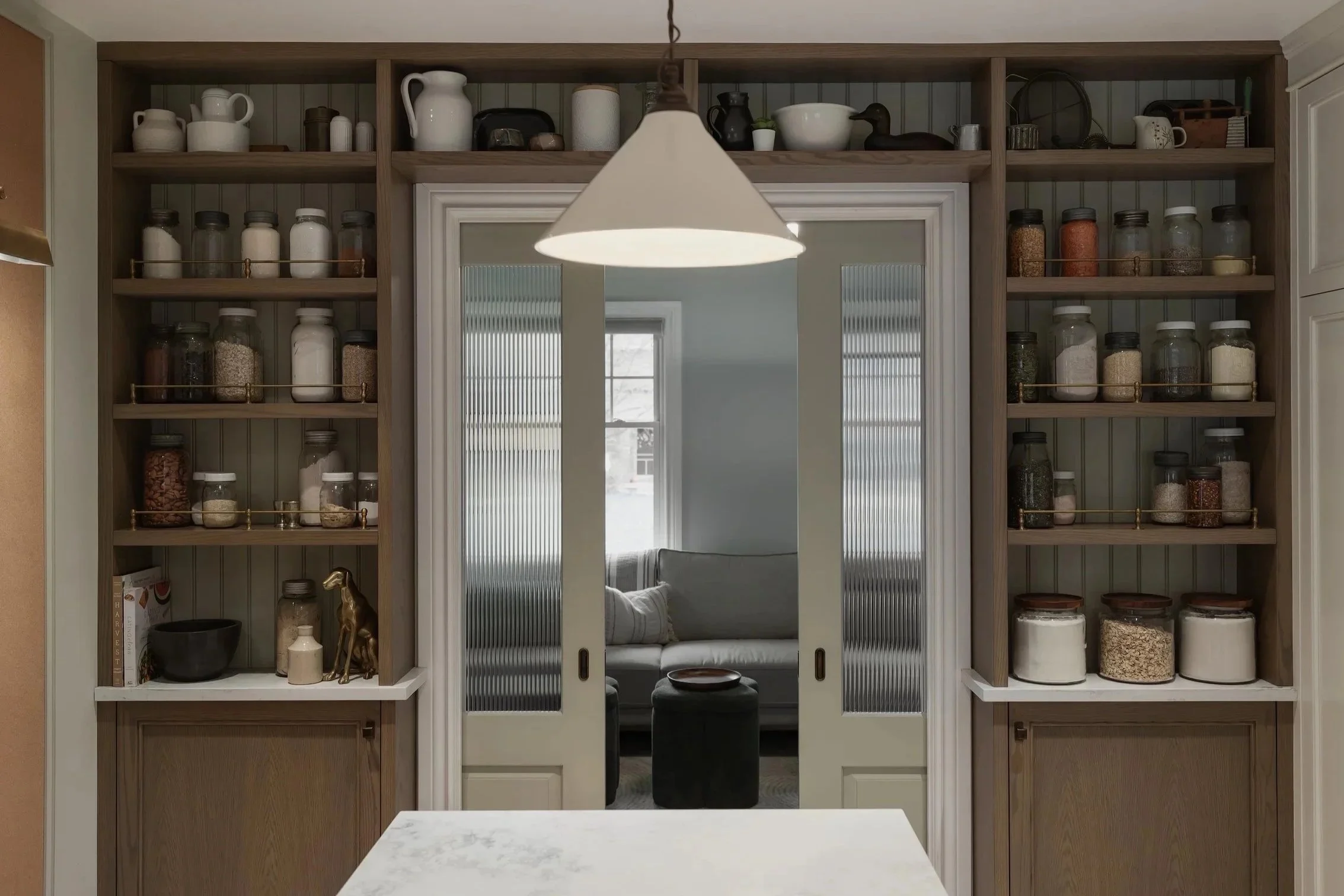 Kitchen with open shelving holding jars of spices and pantry items, sliding doors leading to a living room with a window and sofa, pendant light hanging from ceiling
