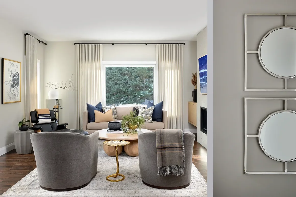 Living room with a white sofa, gray armchairs, and a wooden coffee table, decorated with plants, pillows, and artwork, with large windows and off-white curtains.