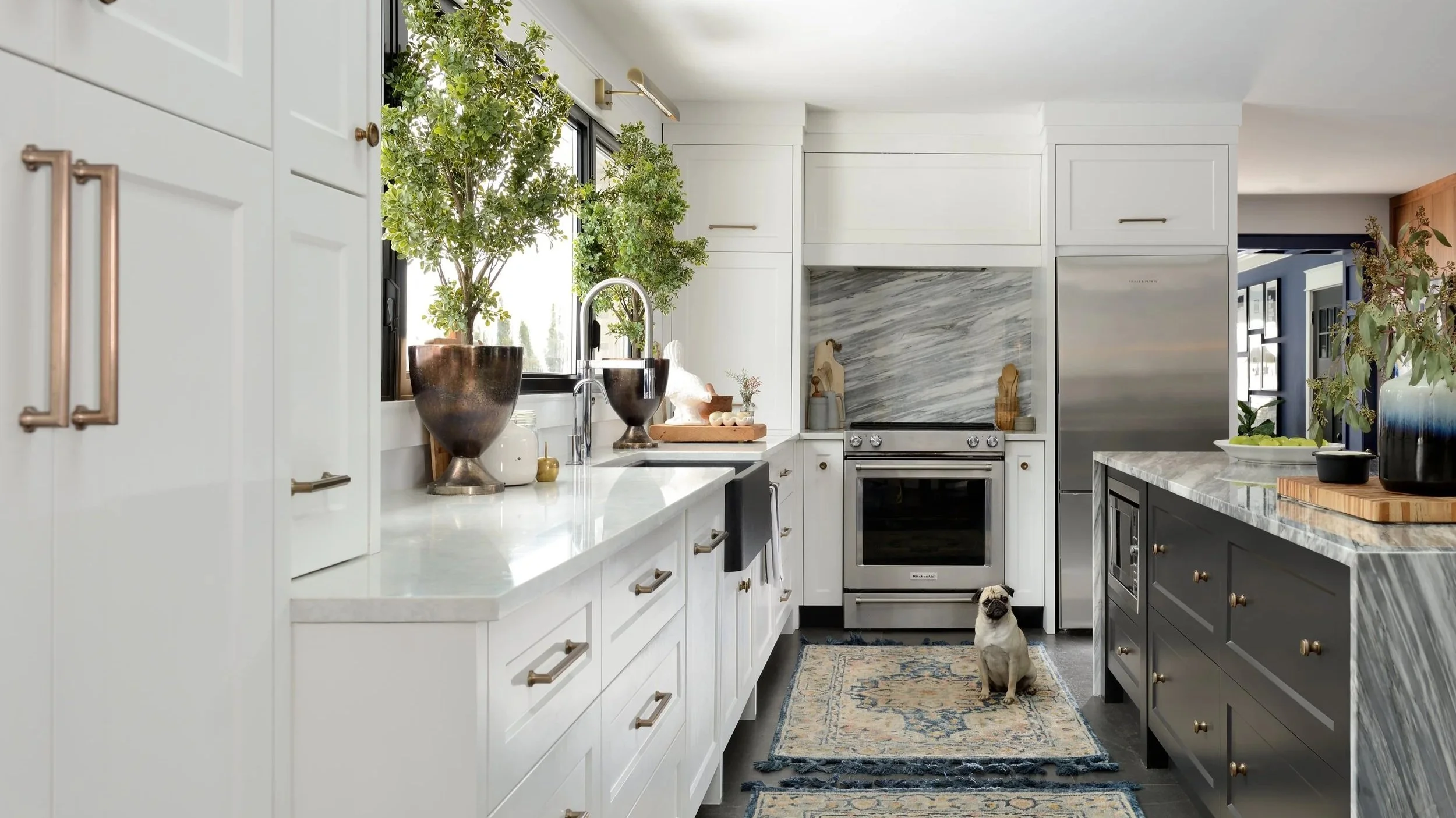 Modern kitchen with white and dark cabinetry, marble countertops, plants, and a pug dog sitting on a patterned rug.