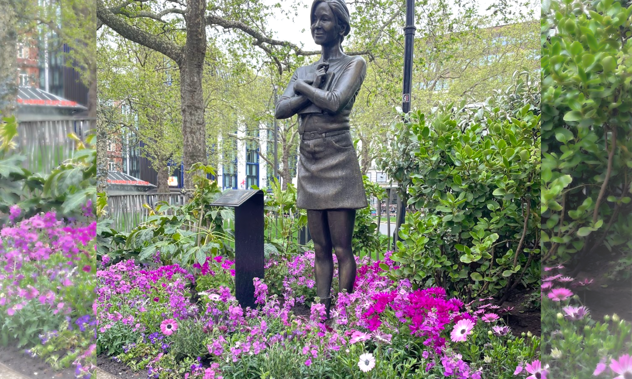 Bronze statue of a woman with short hair, wearing a dress and sweater, holding a book close to her chest, standing among pink and purple flowers and green bushes in a park with trees and buildings in the background.