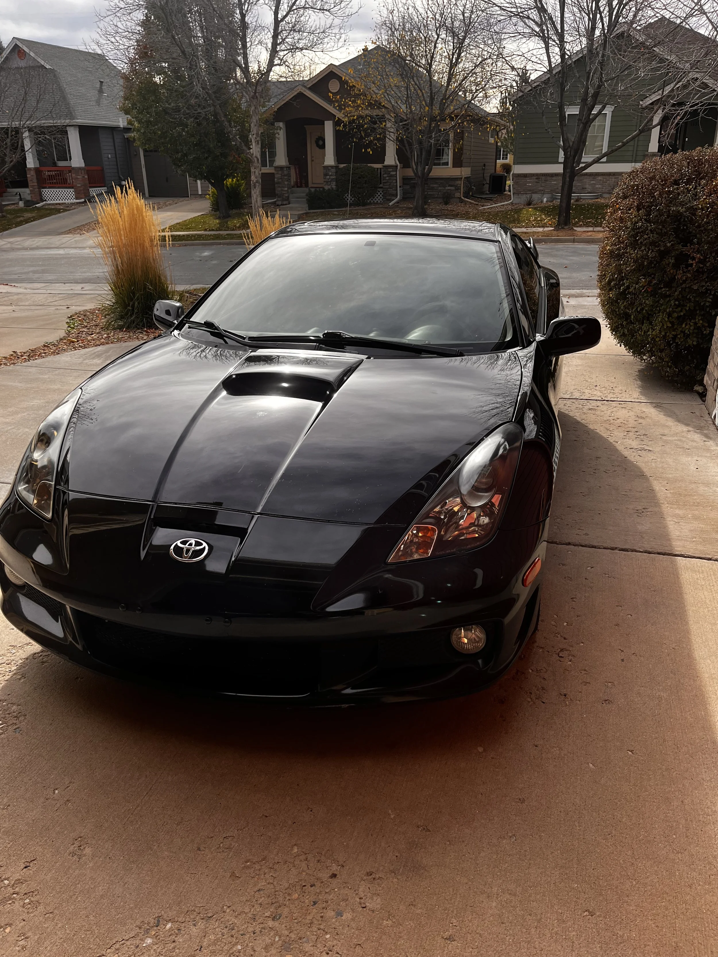 Black Toyota sports car parked in a residential driveway with suburban houses and trees in the background.