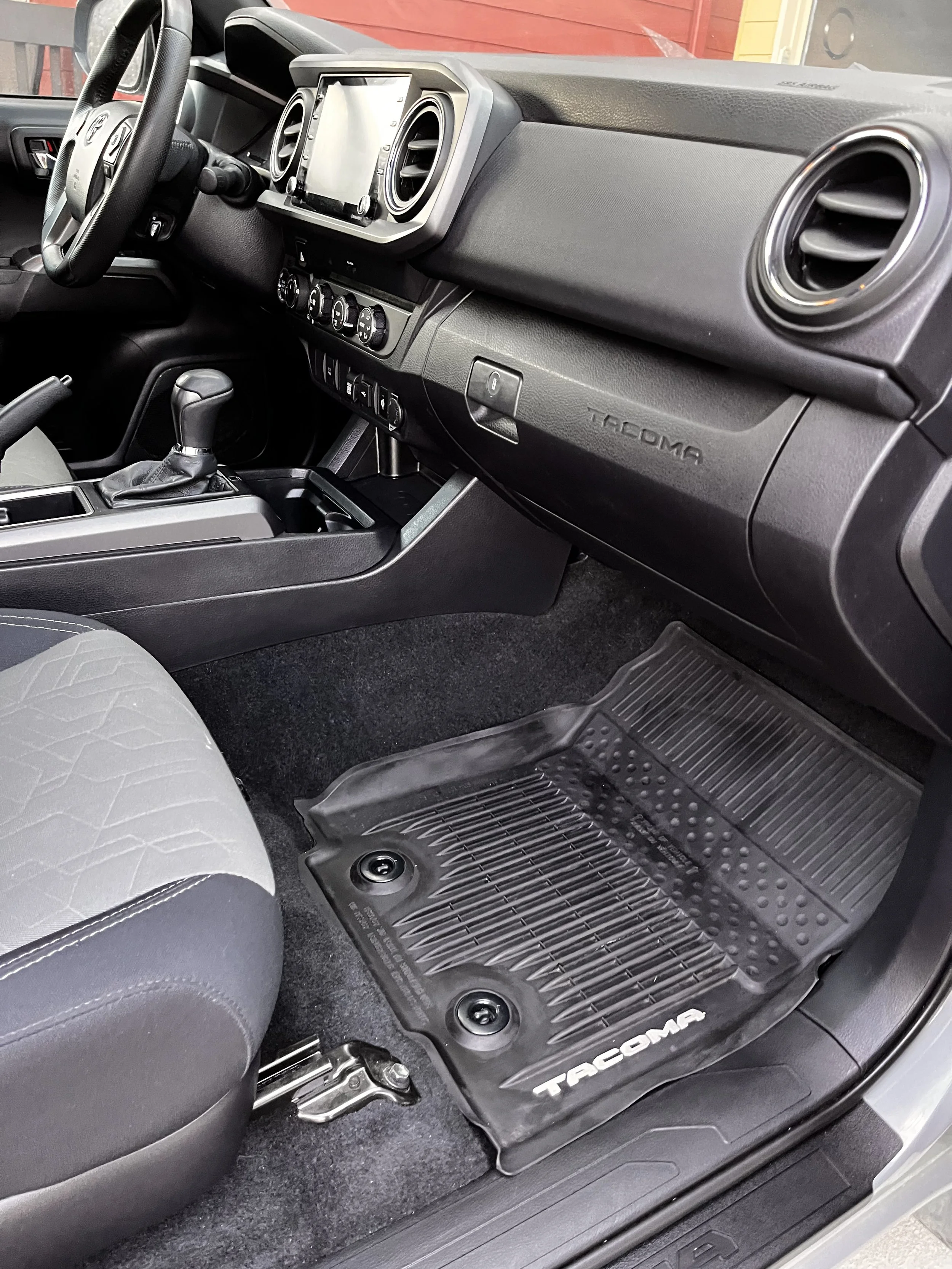 Interior of a Toyota Tacoma truck showing the dashboard, steering wheel, gear shift, and a rubber floor mat with Tacoma branding.