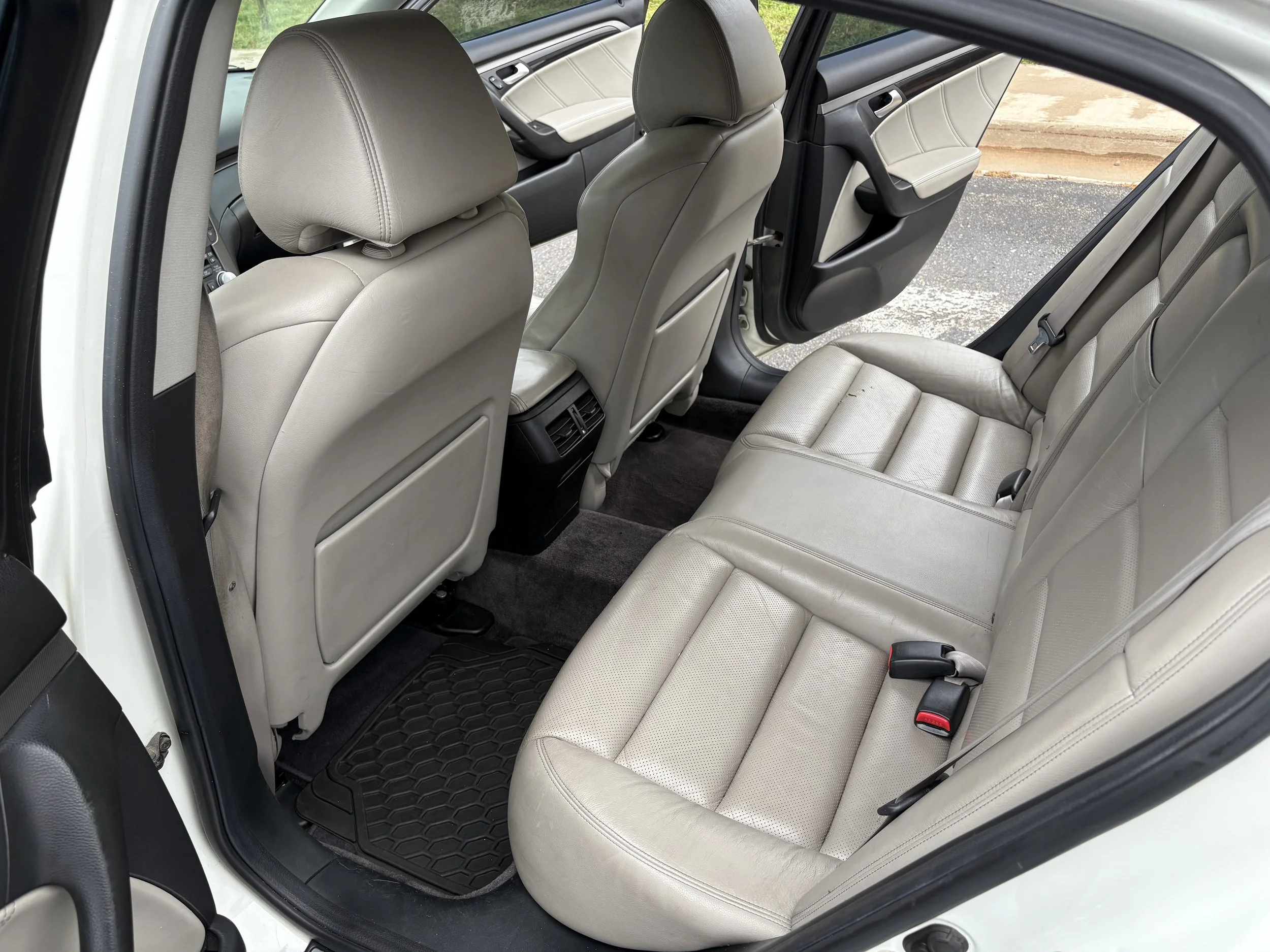Interior view of the back seat of a car with cream-colored leather seats, black floor mats, and beige headrests.