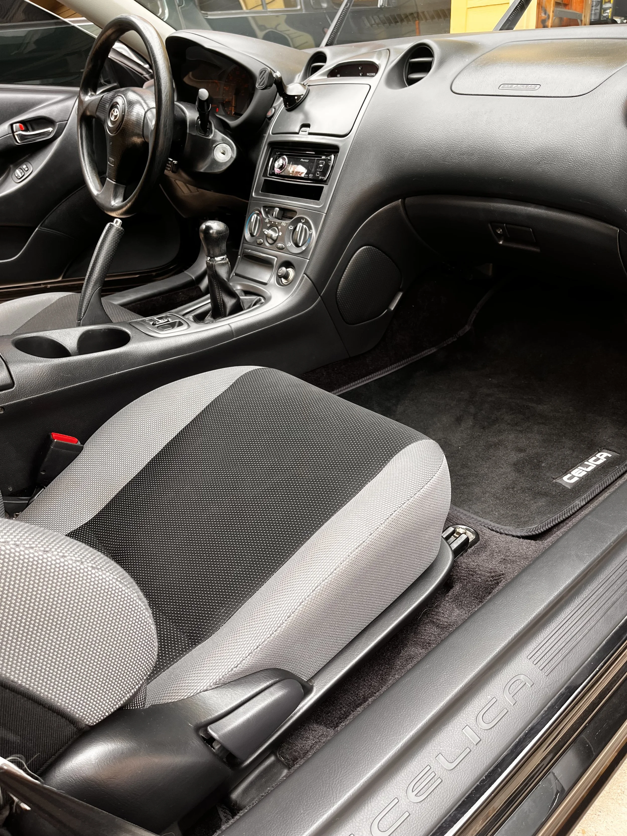Interior of a Toyota Celica, showing the driver's seat, dashboard, steering wheel, gear shift, and front passenger area.