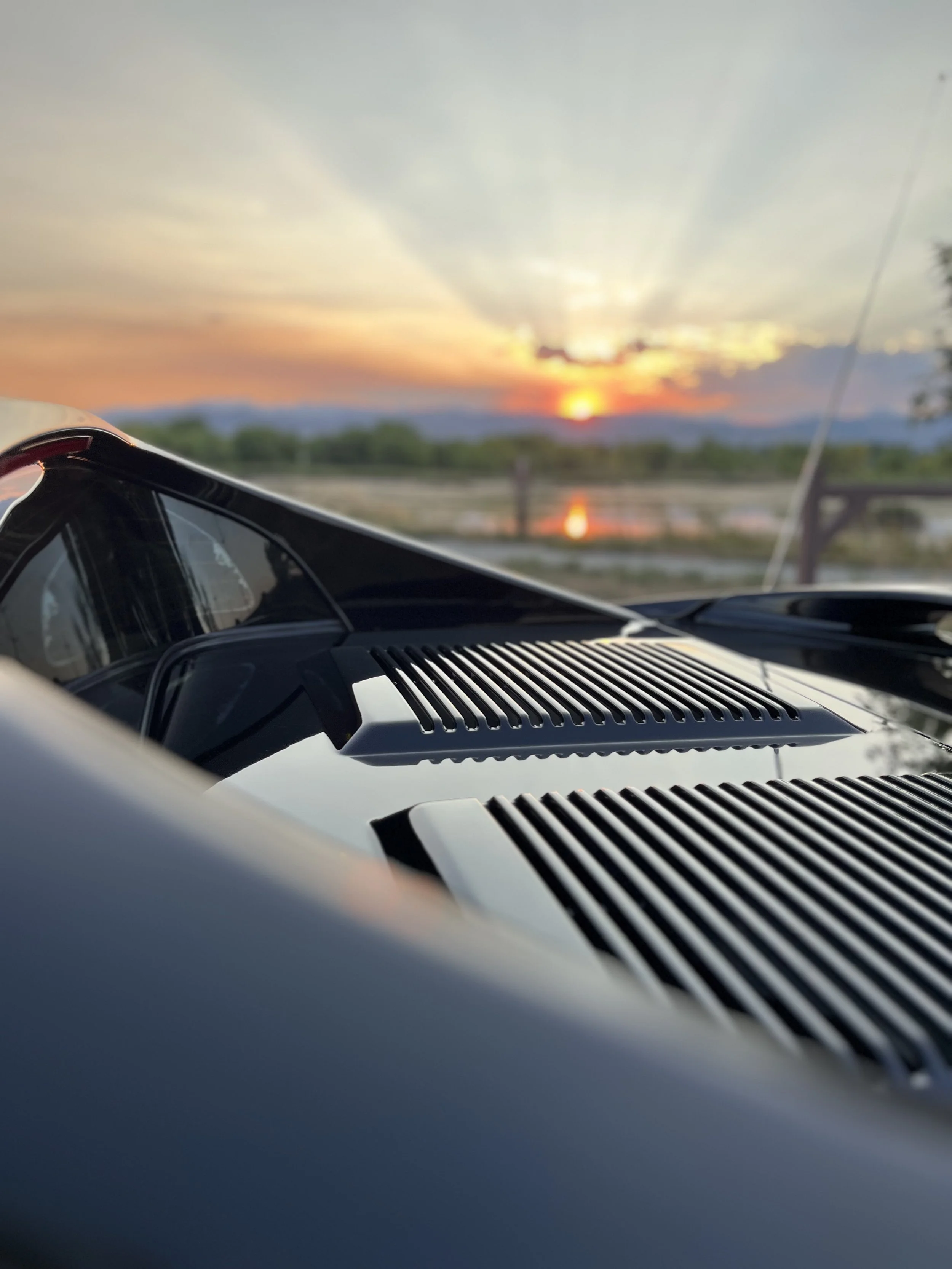 Close-up of a car's hood with vents, sunglasses on top, and a sunset with clouds and a body of water in the background.