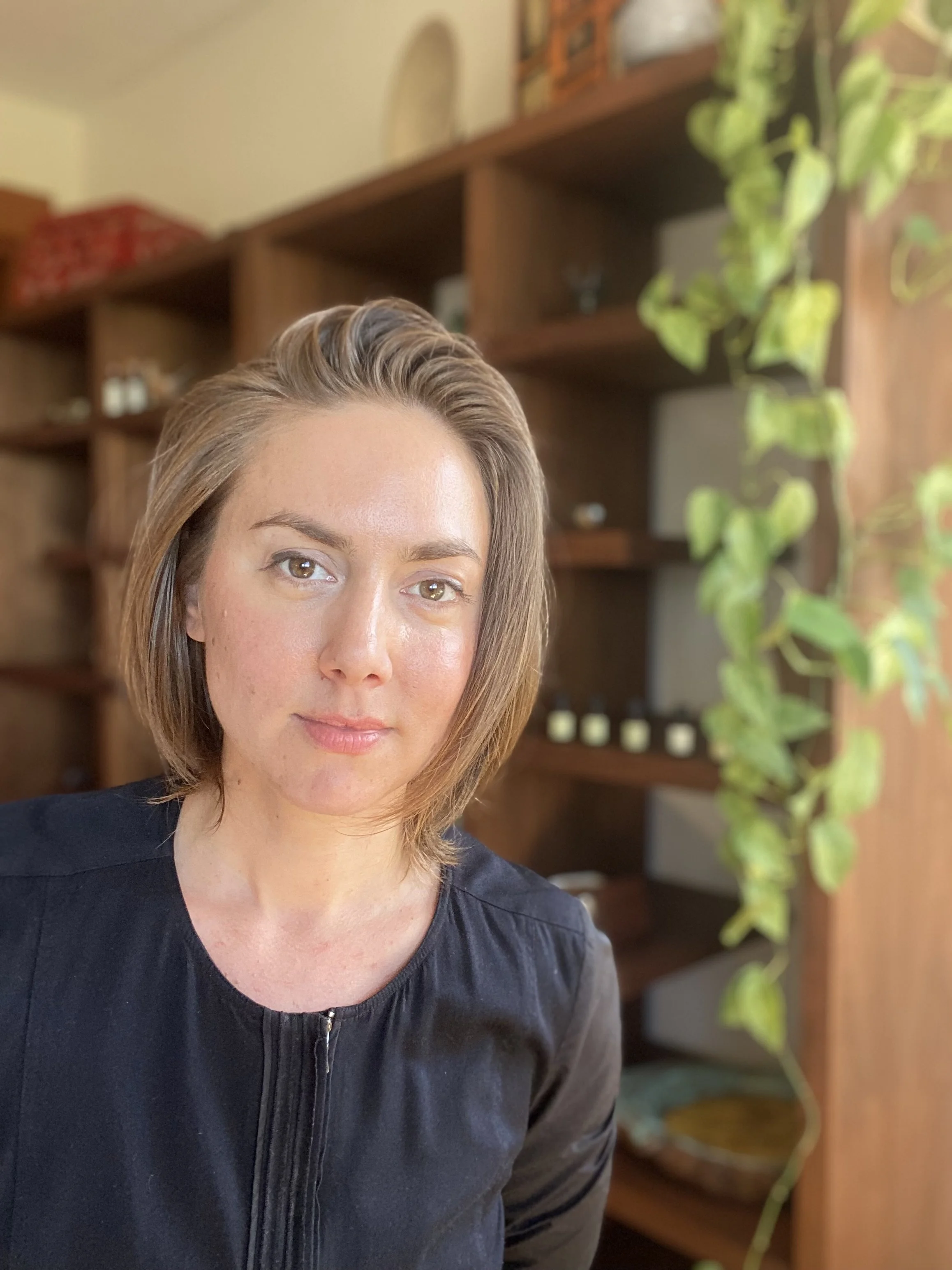 A woman with shoulder-length brown hair and light skin, wearing a black top, is taking a selfie in a room with wooden shelves and a plant in the background.