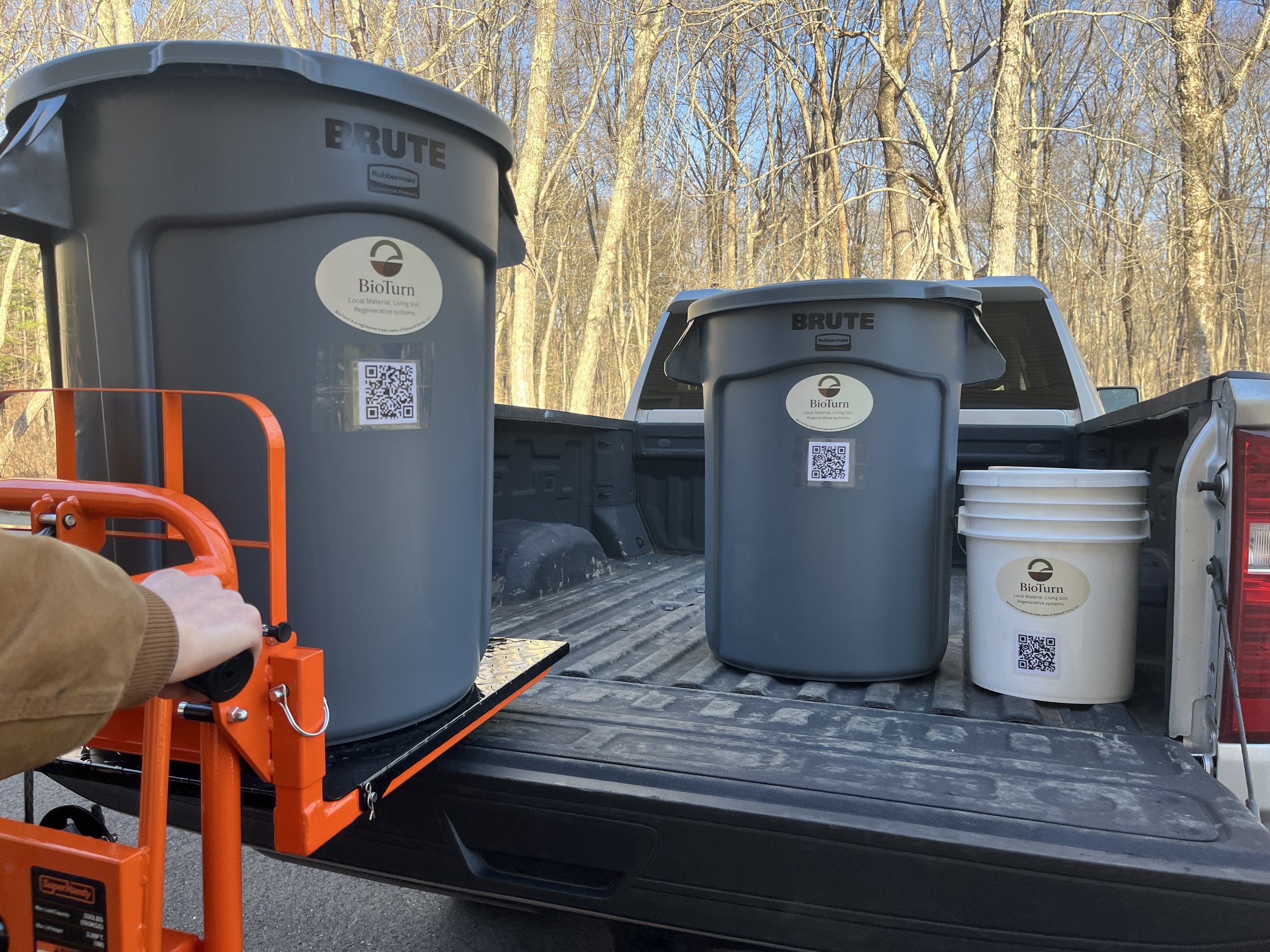 Various gray and white compost bins with QR codes and labels on a pickup truck bed, outdoors with leafless trees in the background.