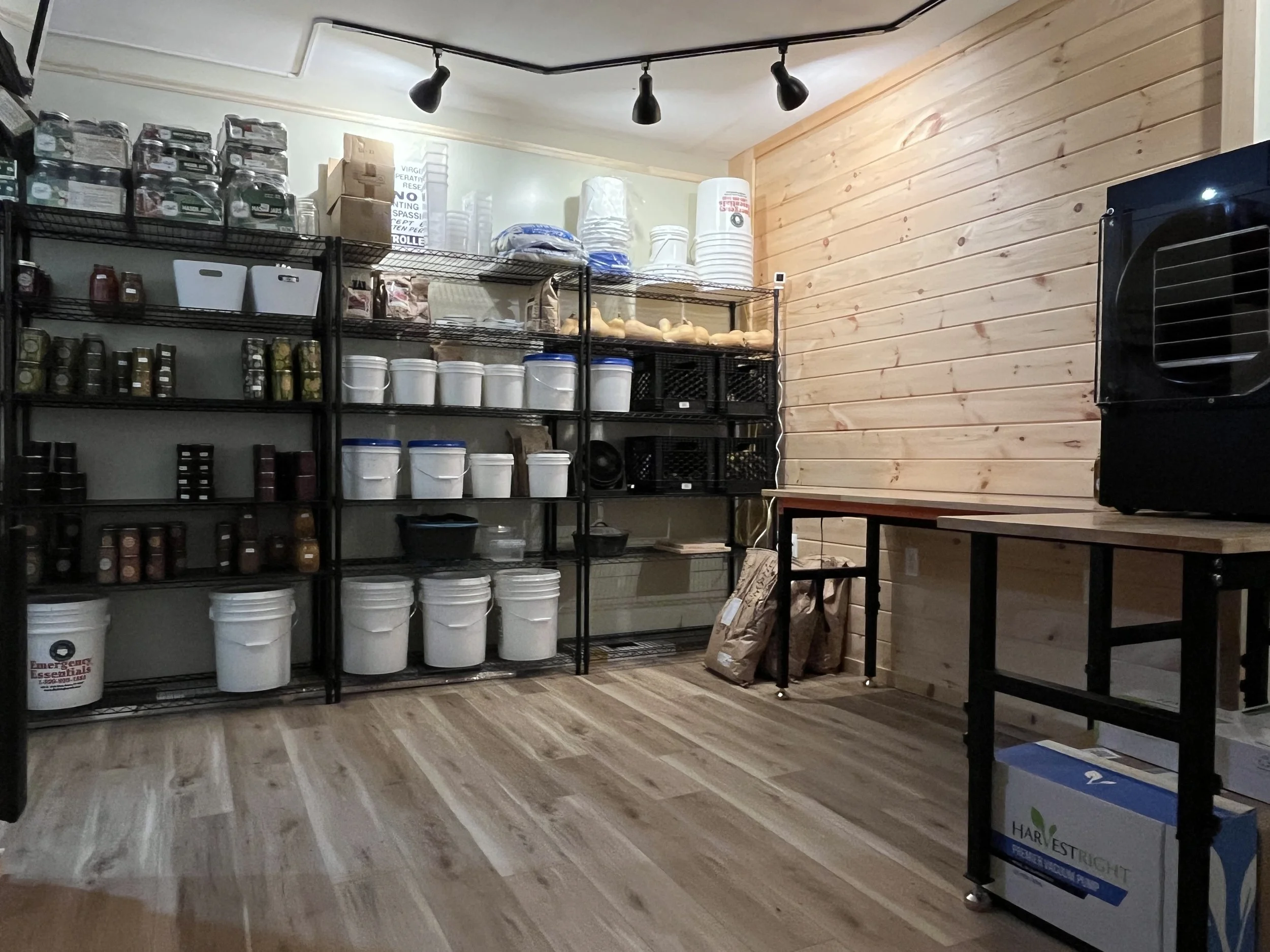 Storage room with black metal shelves filled with white buckets, jars, boxes, and other kitchen supplies, hardwood floor, wooden-paneled wall, black table, and a black appliance on top of another table.