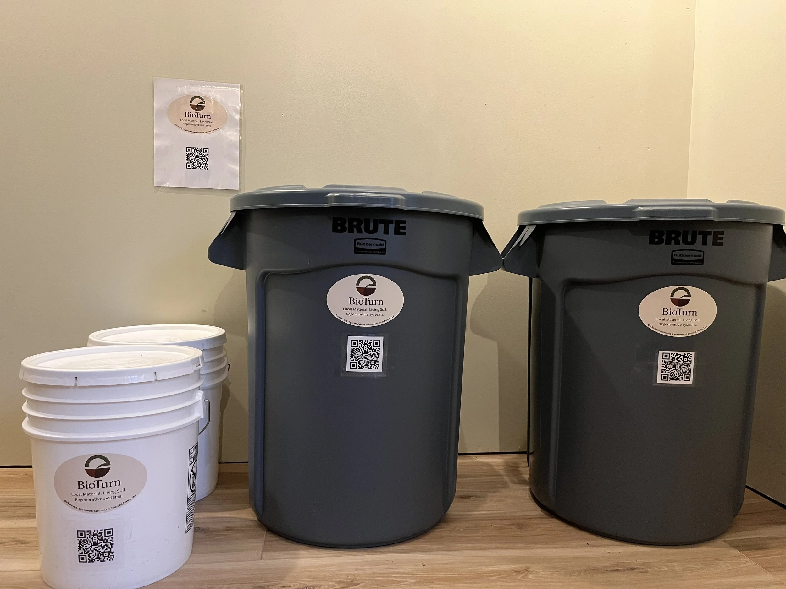 Three white buckets and two gray trash bins labeled BioTurn and BRUTE in a room with a beige wall and wooden floor.