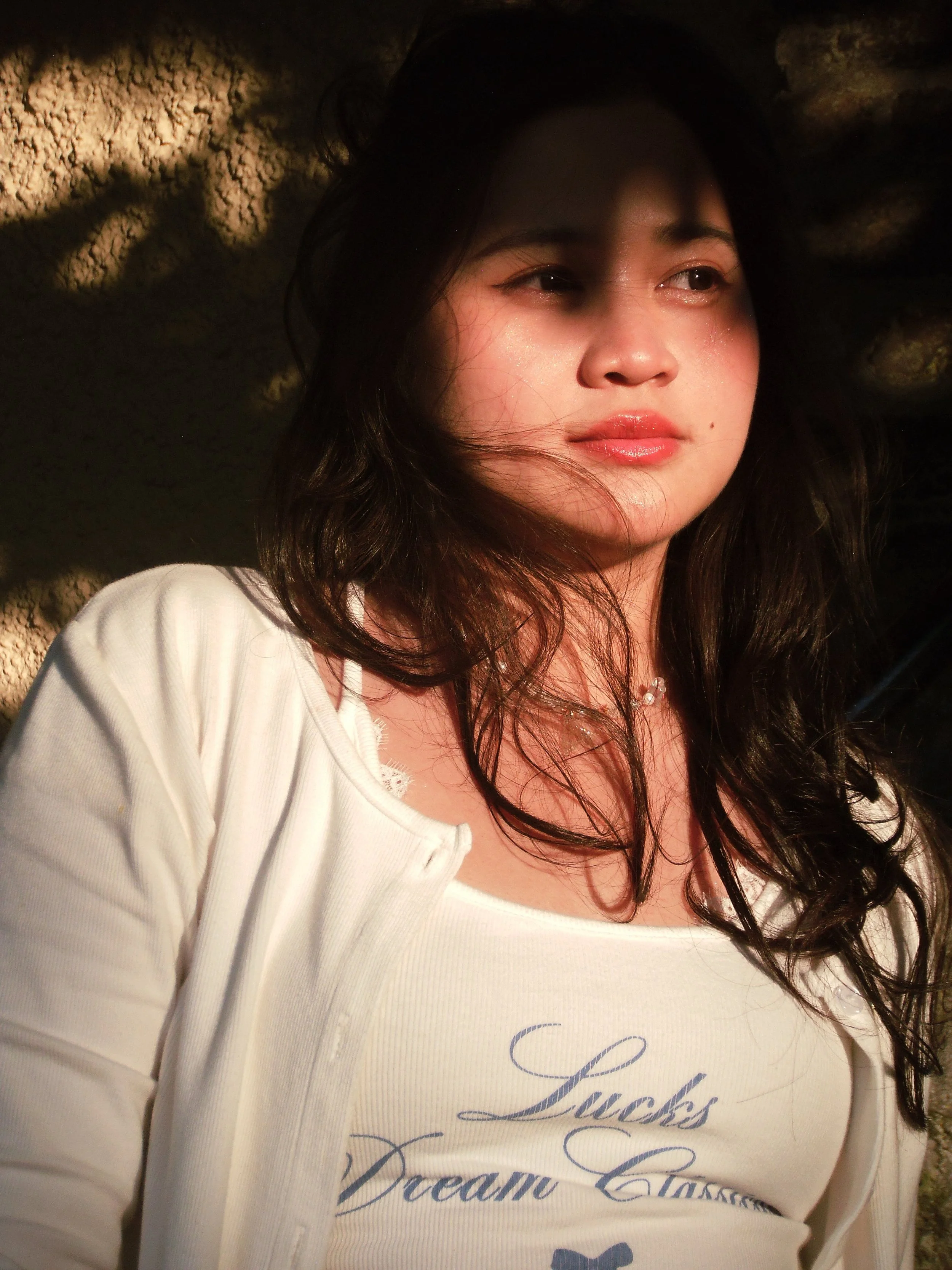 A young woman with long dark hair, wearing a white top and a light jacket, is standing against a rocky background, with sunlight casting shadows across her face.