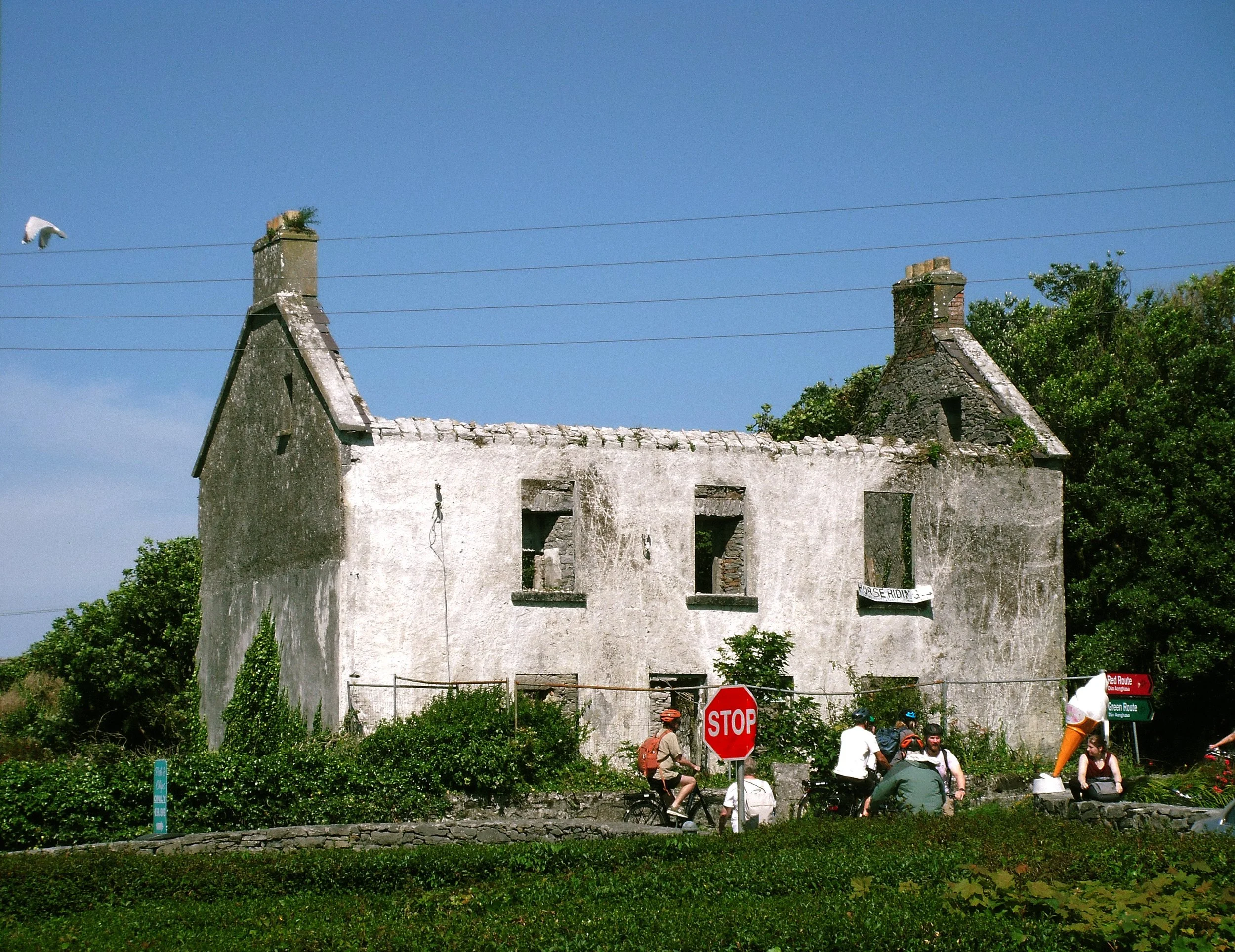 An old, abandoned two-story house with a damaged roof, boarded-up windows, and weathered white walls. Several people are riding bikes and sitting in front of the house, with a stop sign and directional signs nearby. The sky is clear and blue, with gr