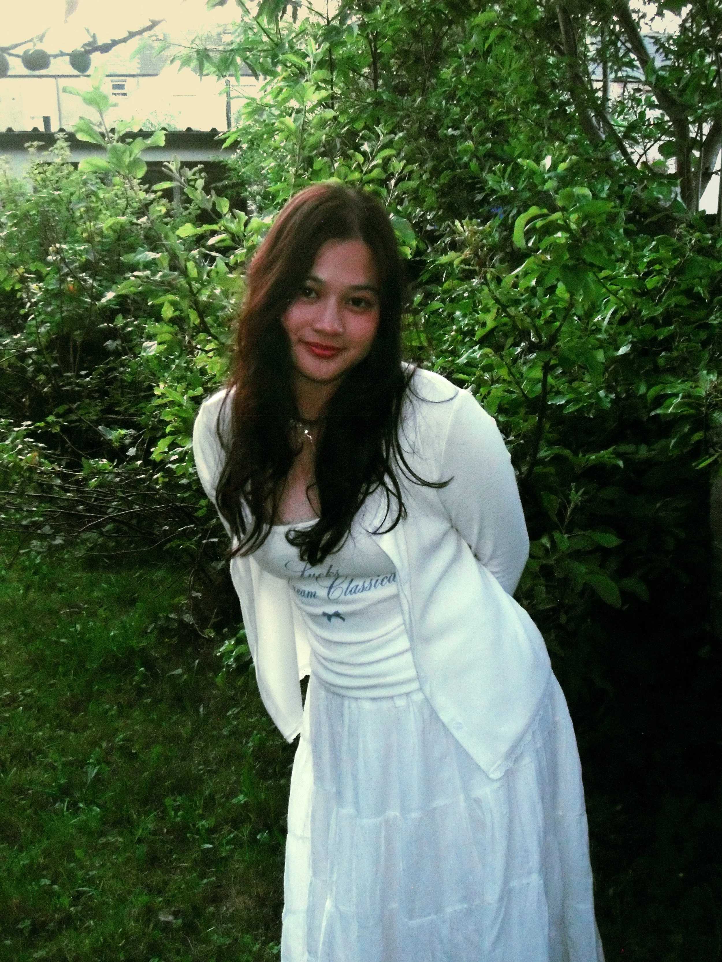 A young woman with long dark hair wearing a white jacket and white dress standing outdoors in front of green bushes.