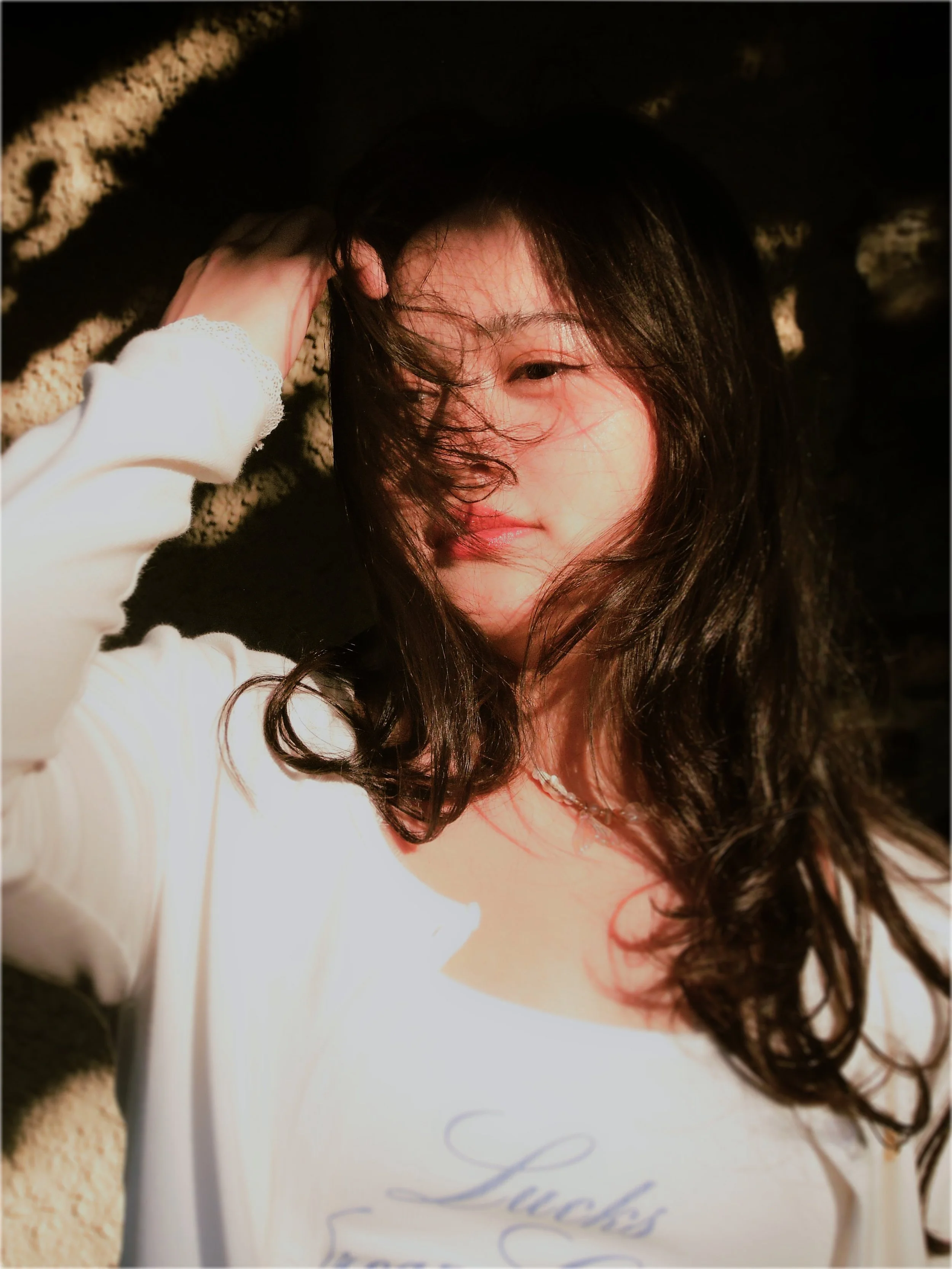 A woman with long dark hair lying on sand at the beach, wearing a white shirt. She has a subtle smile, and her hair is blowing across her face, with her hand resting on her head.