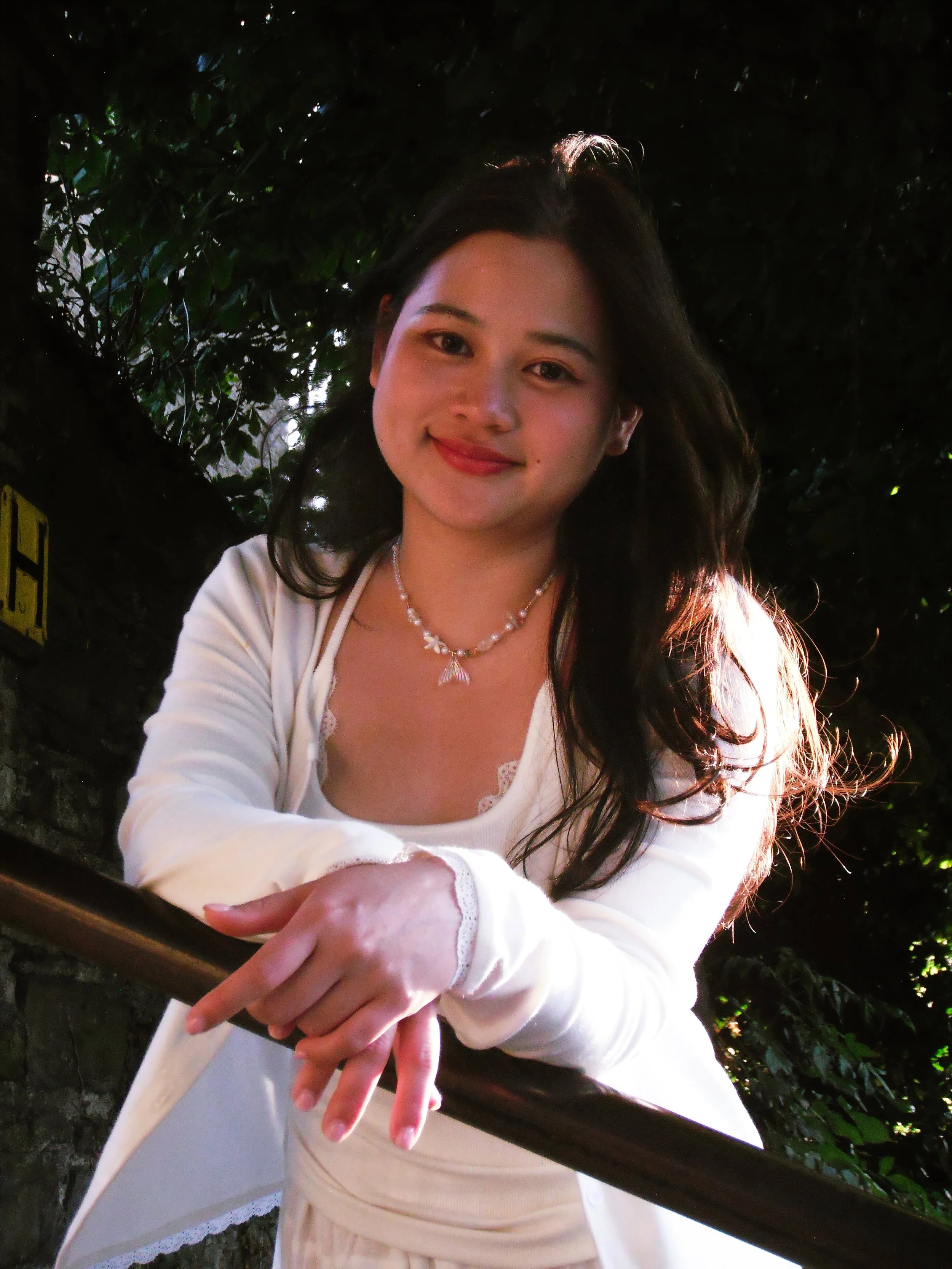 A young woman with long dark hair, smiling, wearing a white outfit and jewelry, leaning on a railing outdoors with trees in the background.