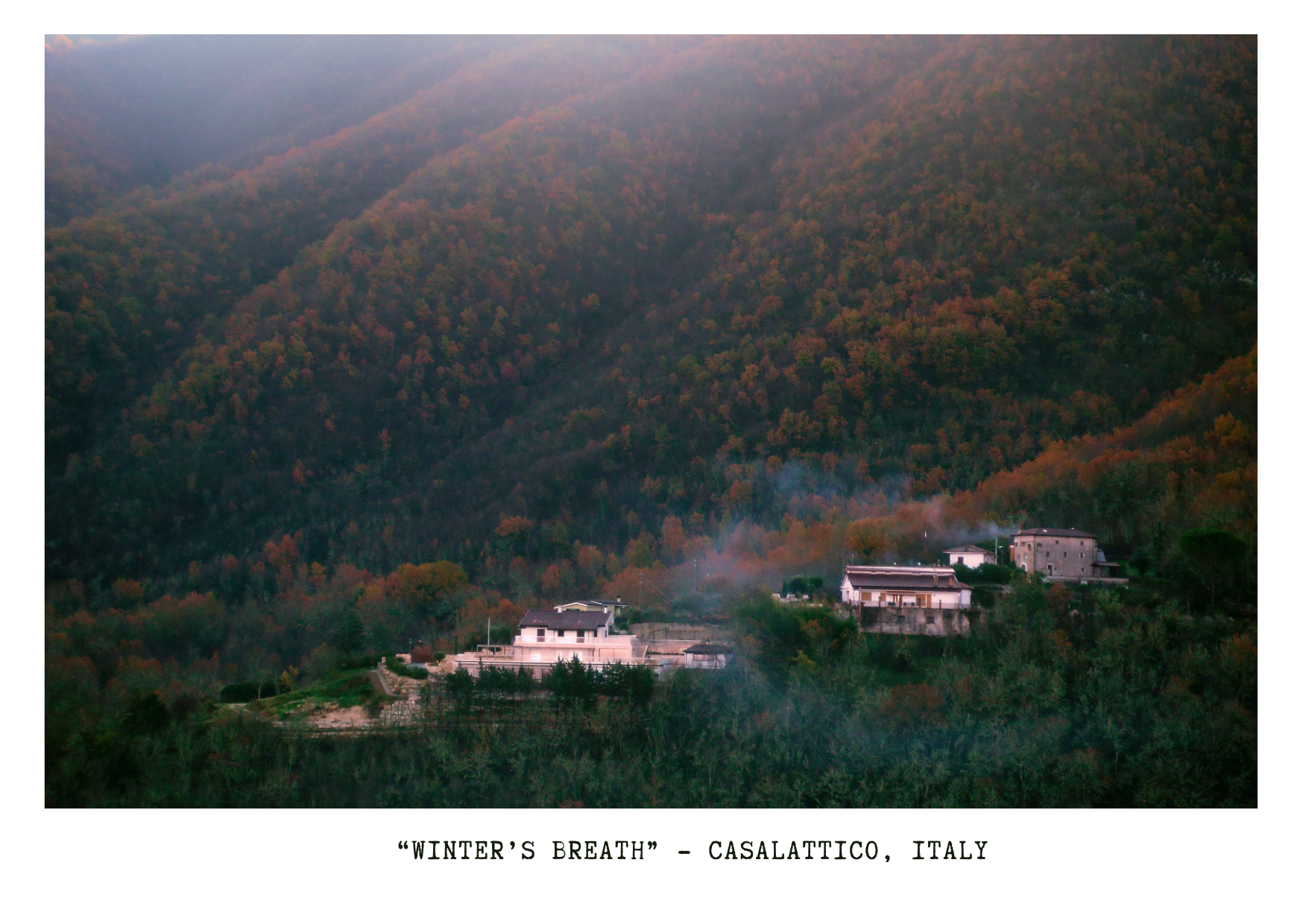 Scenic view of houses nestled on a hillside with autumn-colored forested mountains in the background, with hot smoke rising from chimneys, captioned 'Winter's Breath' - Casalattico, Italy.