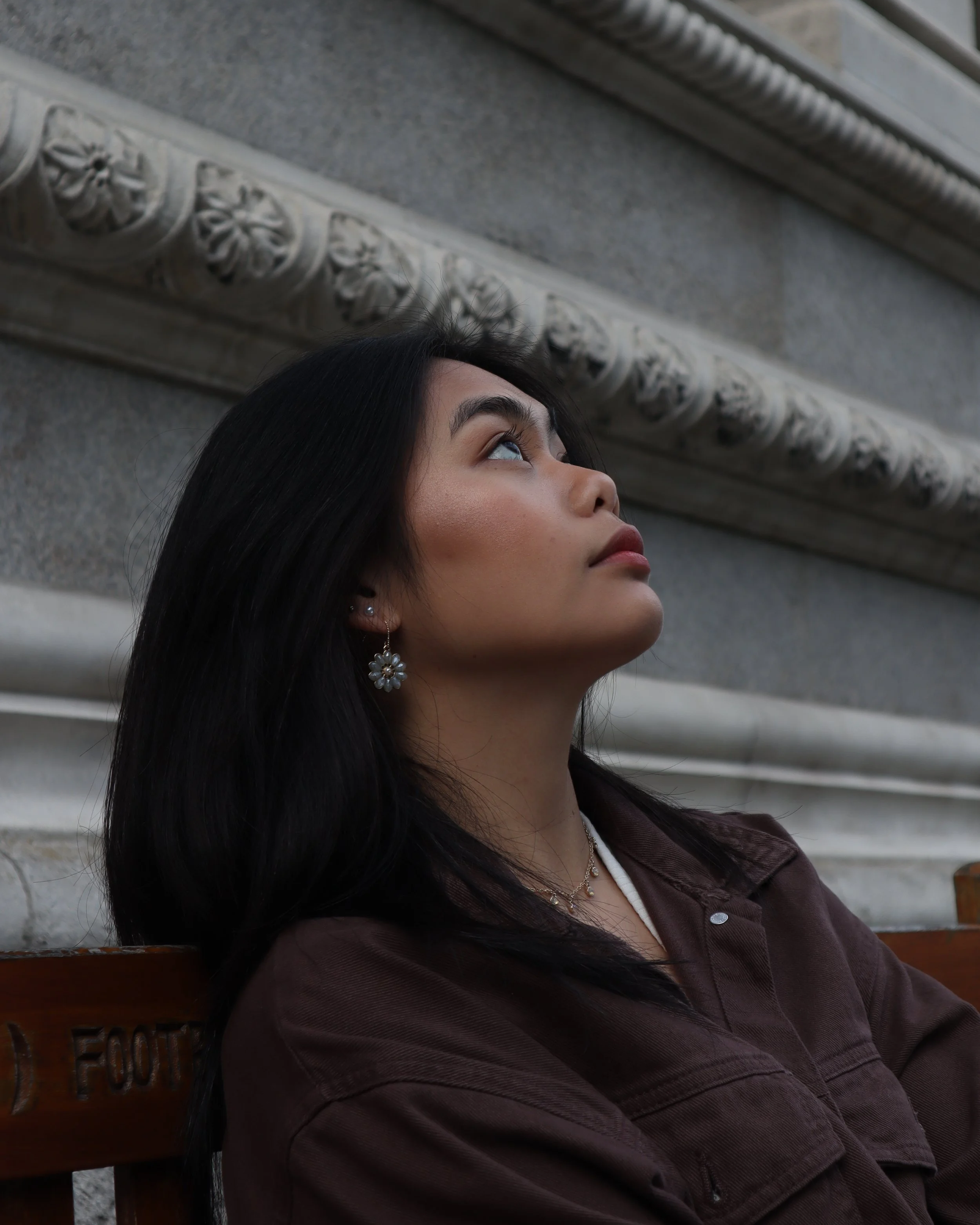 A young woman with black hair and earrings sitting on a bench, looking up at the ornate architectural detail on a building's wall.