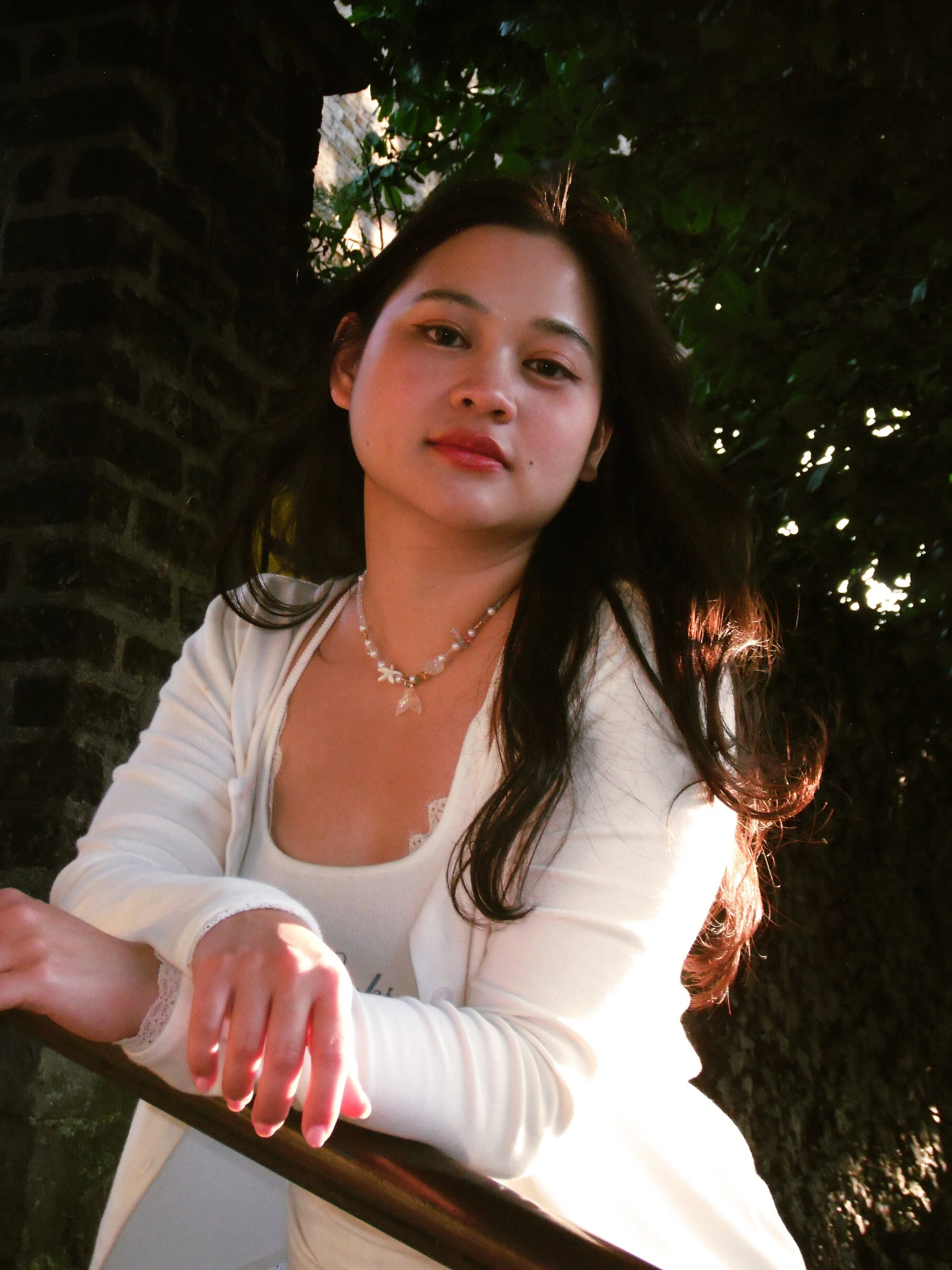 A woman with dark hair wearing a white top and necklace, leaning on a railing outdoors near a tree.