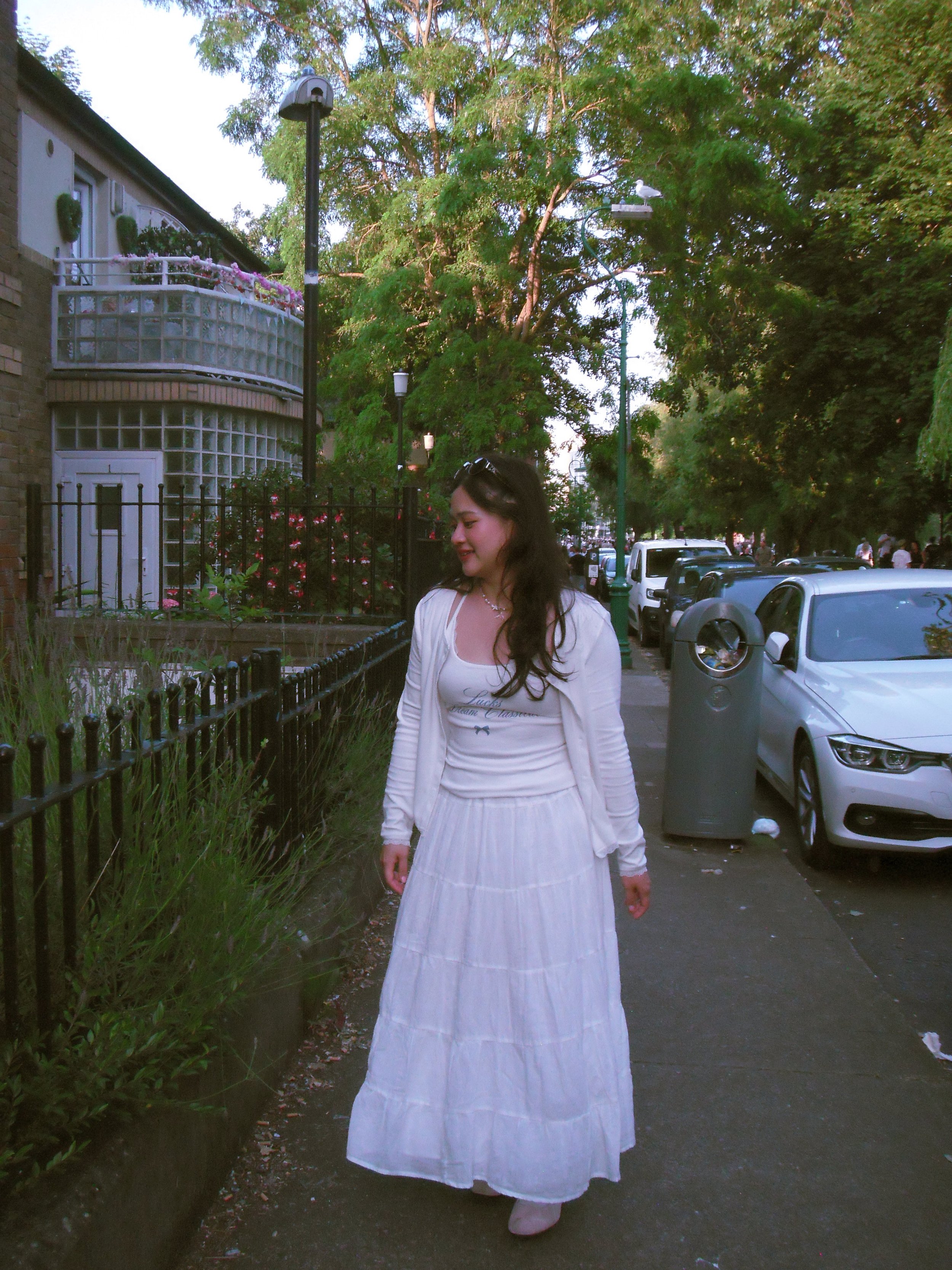 A woman in a white outfit walking on a sidewalk lined with greenery and parked cars, with trees, street lamps, and a building with a balcony in the background.